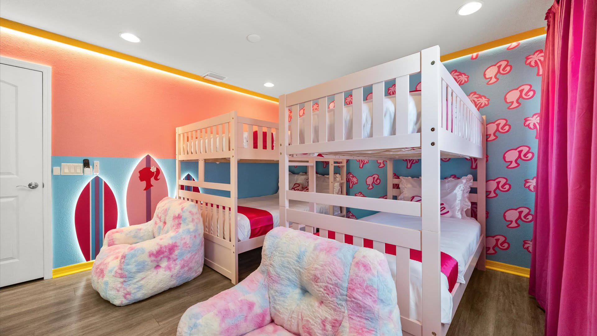 Vibrant Barbie-themed bedroom with two white bunk beds, pink bedding, and two tie-dye bean bag chairs. Walls feature a mix of pink and blue with surfboard and Barbie head graphics, and bright pink curtains.