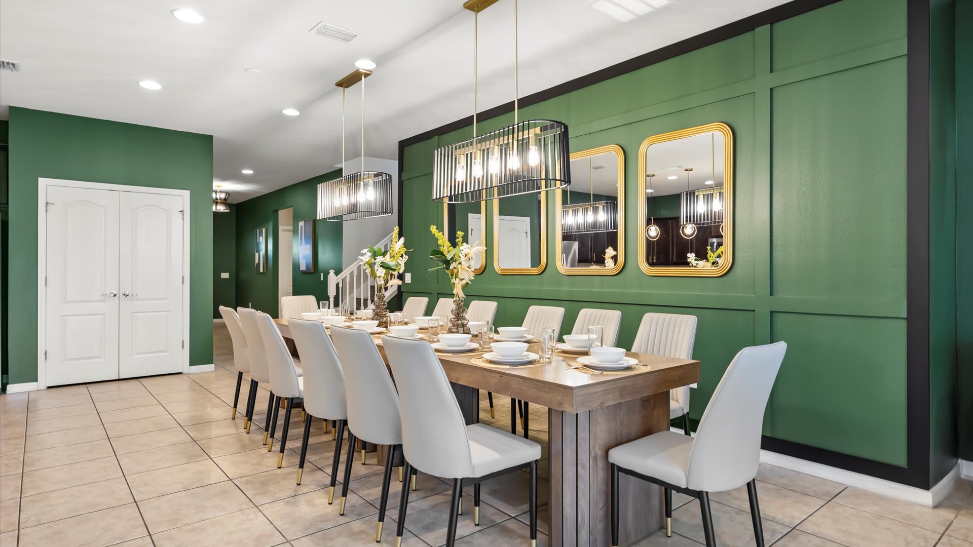 A spacious dining room featuring a long wooden table with seating for twelve, set against a dark green paneled accent wall. Two modern chandeliers illuminate the space, complemented by gold-framed mirrors and tile flooring.