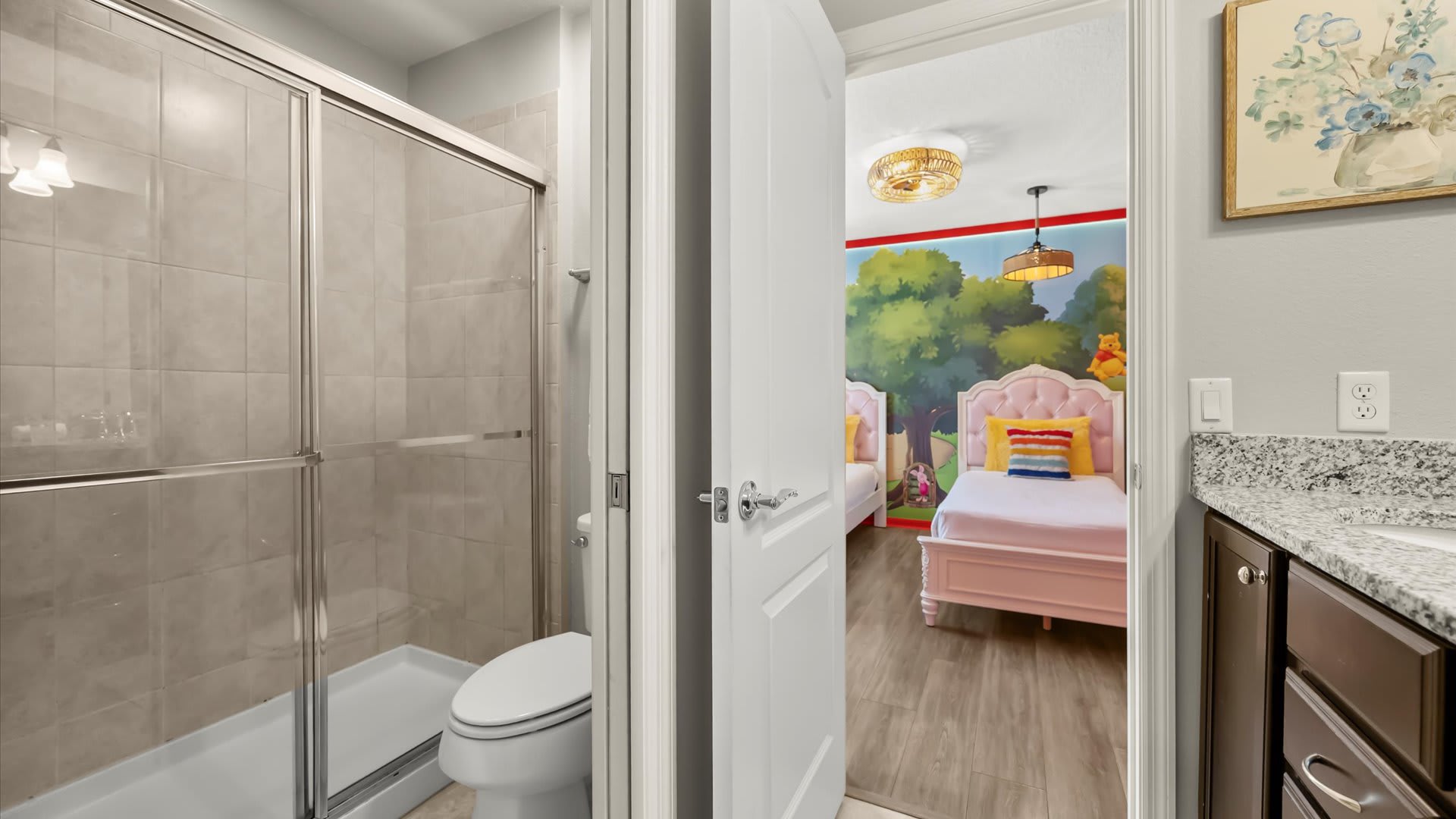 A modern bathroom featuring a glass-enclosed shower, a white toilet, and a vanity with a granite countertop, with a partial view into a pink-themed bedroom.