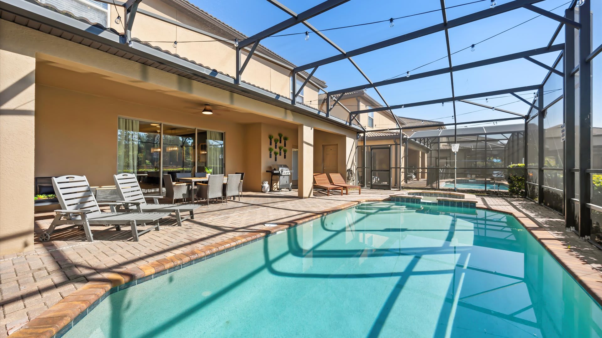 A wide view of the screened-in outdoor pool area, showcasing the clear blue swimming pool and hot tub. The spacious brick paver deck includes a covered patio with outdoor seating and a grill, all under a bright sky.