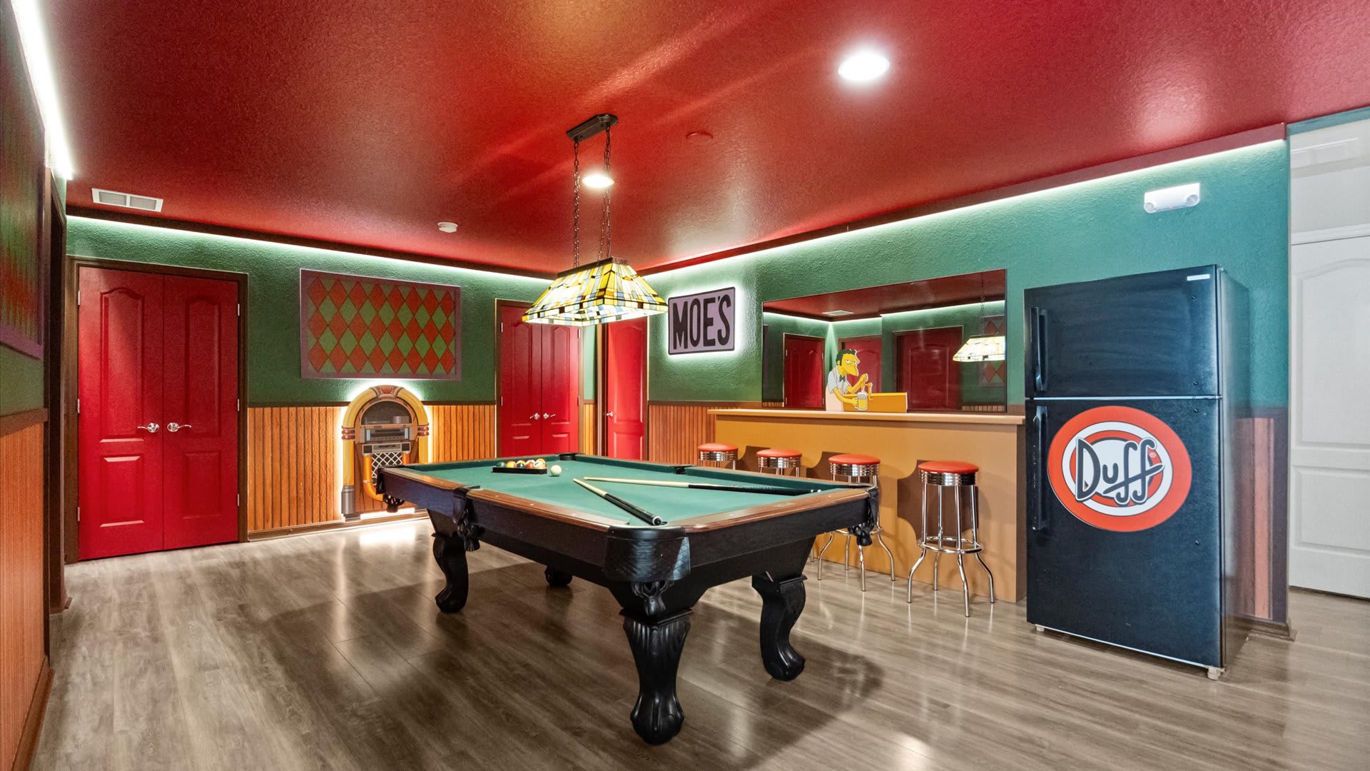 A themed game room, reminiscent of Moe's Tavern, features a pool table under a stained-glass lamp, a bar with stools, and a "Duff" branded refrigerator. The room has red ceilings, teal walls, and wood paneling, with red doors and a jukebox.
