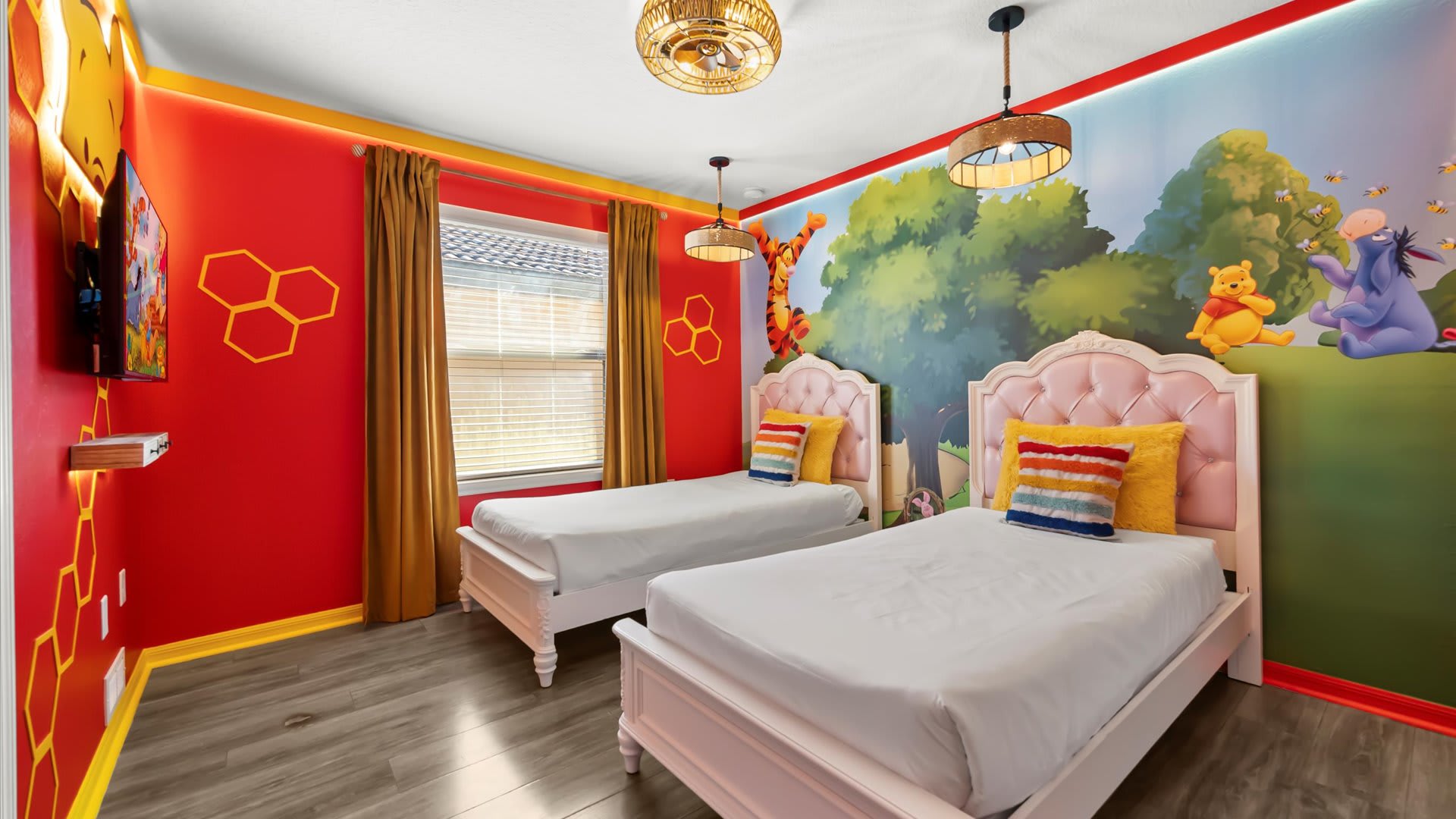 A vibrant Winnie the Pooh themed bedroom features two pink twin beds with white bedding, colorful pillows, and a large mural depicting characters from the Hundred Acre Wood. Red walls with honeycomb accents and unique pendant lights complete the playful design.