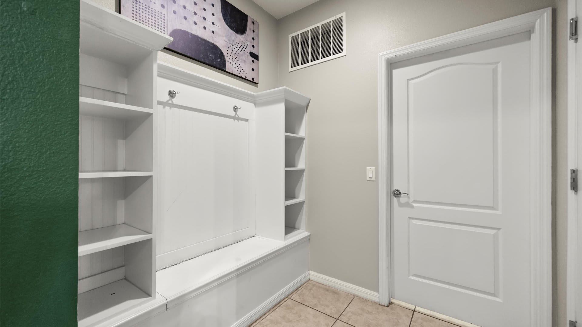 A functional mudroom area with white built-in shelving, coat hooks, and a bench, situated next to a white door and light-colored tiled flooring.