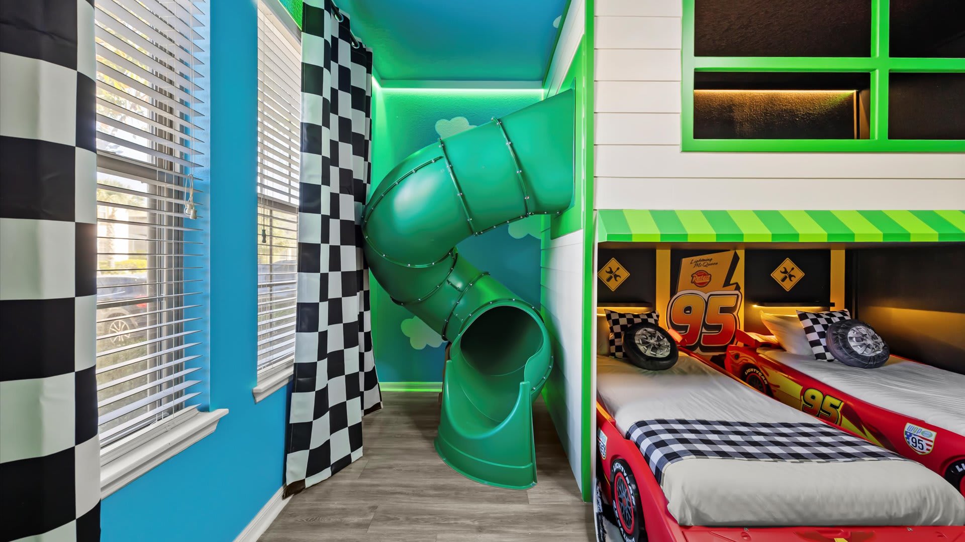 A fun green spiral slide integrated into the Cars-themed bedroom, positioned next to a window with checkerboard curtains and light blue walls.
