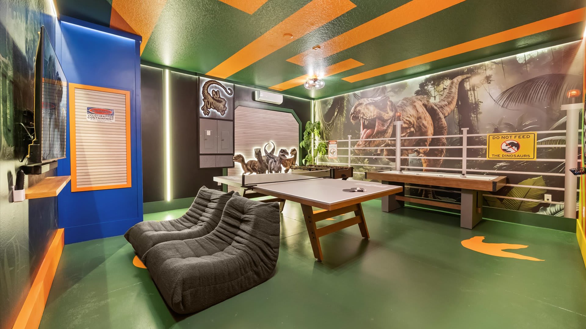 A vibrant dinosaur-themed game room features a green floor with orange accents, a ping-pong table, and large dinosaur murals on the walls. Blue and orange wall panels, bean bag chairs, and neon lighting complete the playful atmosphere.