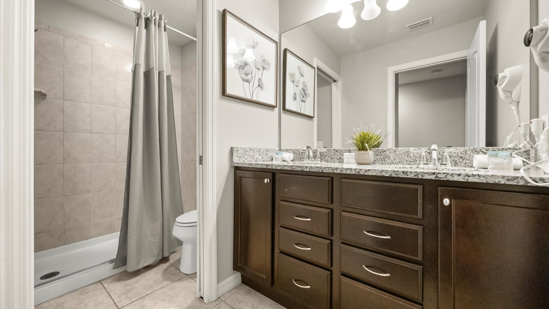 This bathroom features a dark wood double vanity with a granite countertop and two sinks, a large mirror, and a shower/tub combo with a gray shower curtain. The light gray walls and tiled floors maintain a clean and functional design.