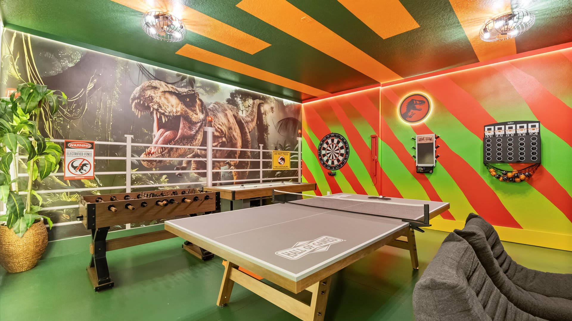 A vibrant Jurassic Park-themed game room featuring a ping pong table, foosball table, dartboard, and air hockey. The space boasts bold dinosaur murals, a striped ceiling, and a bean bag chair.