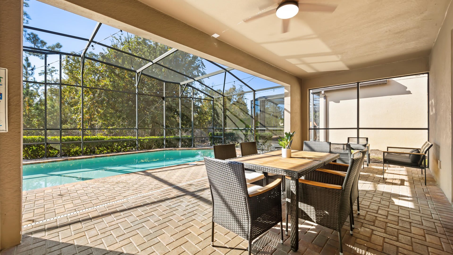 A covered outdoor patio features a large dining table with wicker chairs, overlooking a screened-in swimming pool and lush green trees. The brick paver floor and ceiling fan provide a comfortable outdoor dining experience.