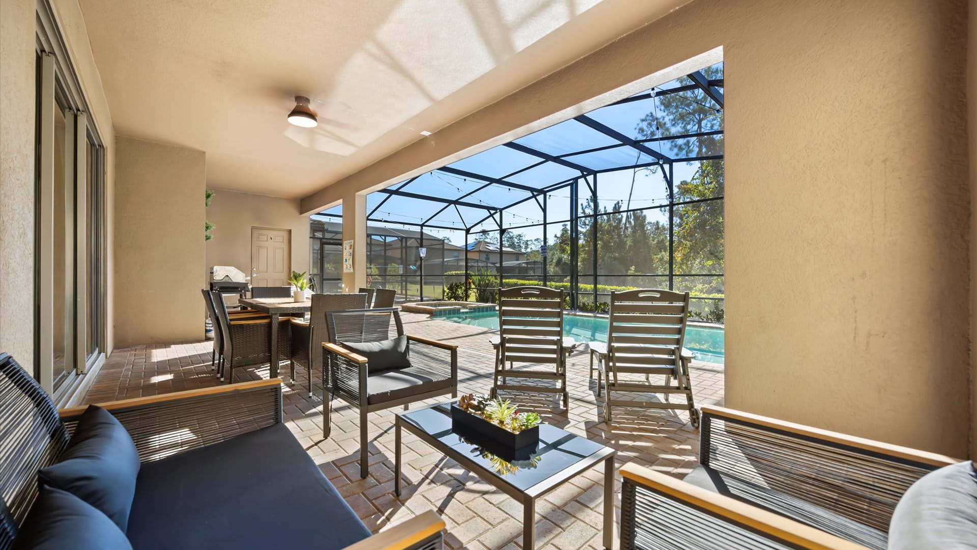 A covered outdoor patio area features comfortable lounge seating with dark cushions and a modern coffee table, positioned on a brick paver deck. A dining table and the screened pool are visible in the background.