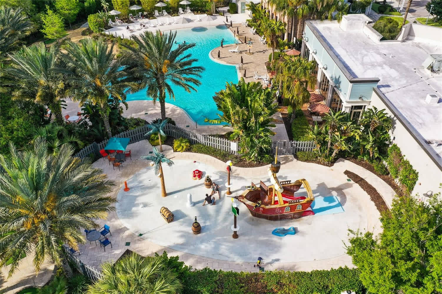 An aerial view of a vibrant resort pool area, featuring a large lagoon-style swimming pool with blue water and a separate children's splash pad with a pirate ship play structure. Palm trees and lounge chairs surround the pools, with a building visible in the background.