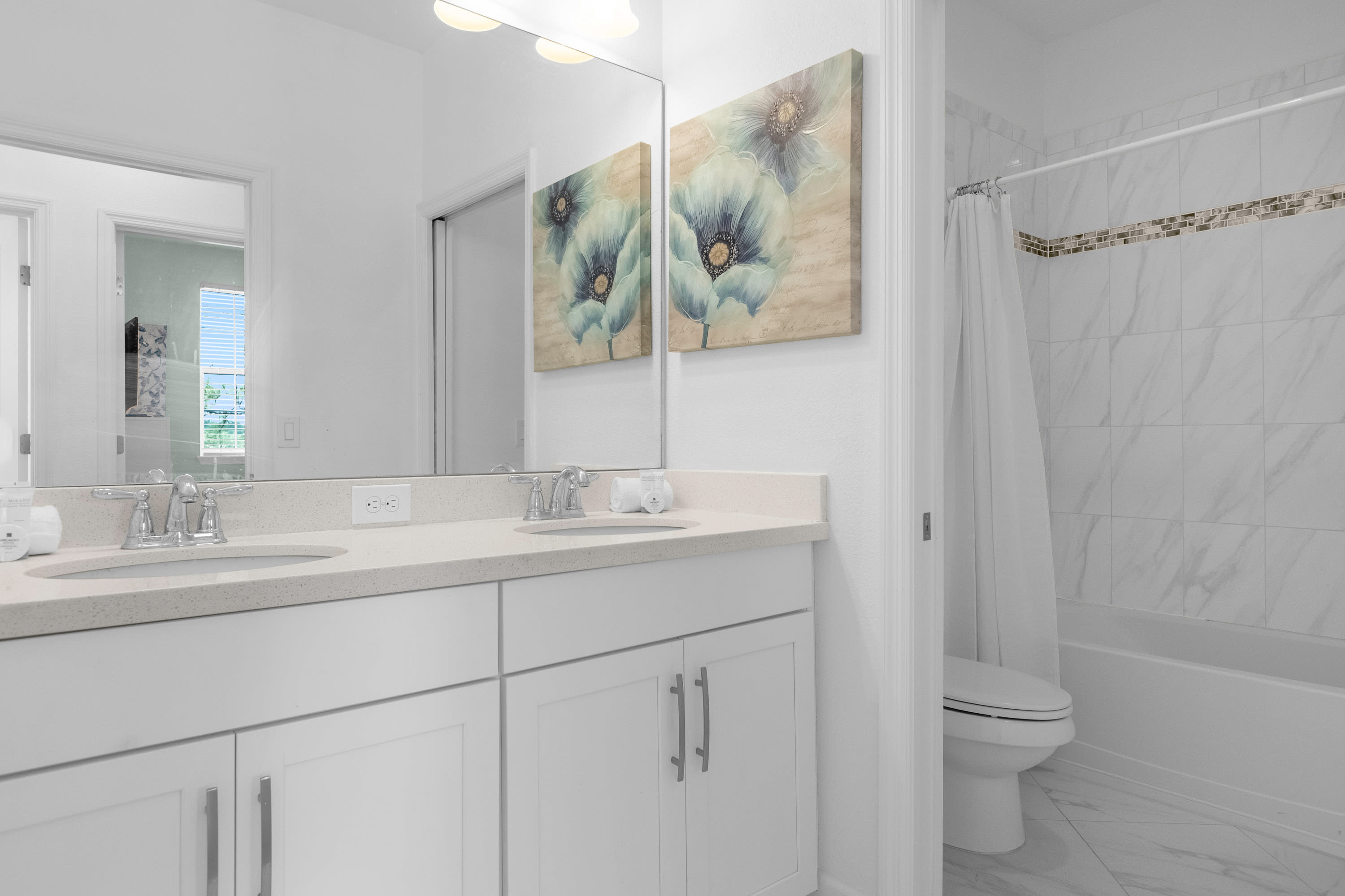 View of the bathroom's double vanity with light countertops and chrome fixtures, reflecting a floral artwork in the mirror. To the right, a white toilet is visible next to the shower-tub combo, maintaining a clean and bright aesthetic.