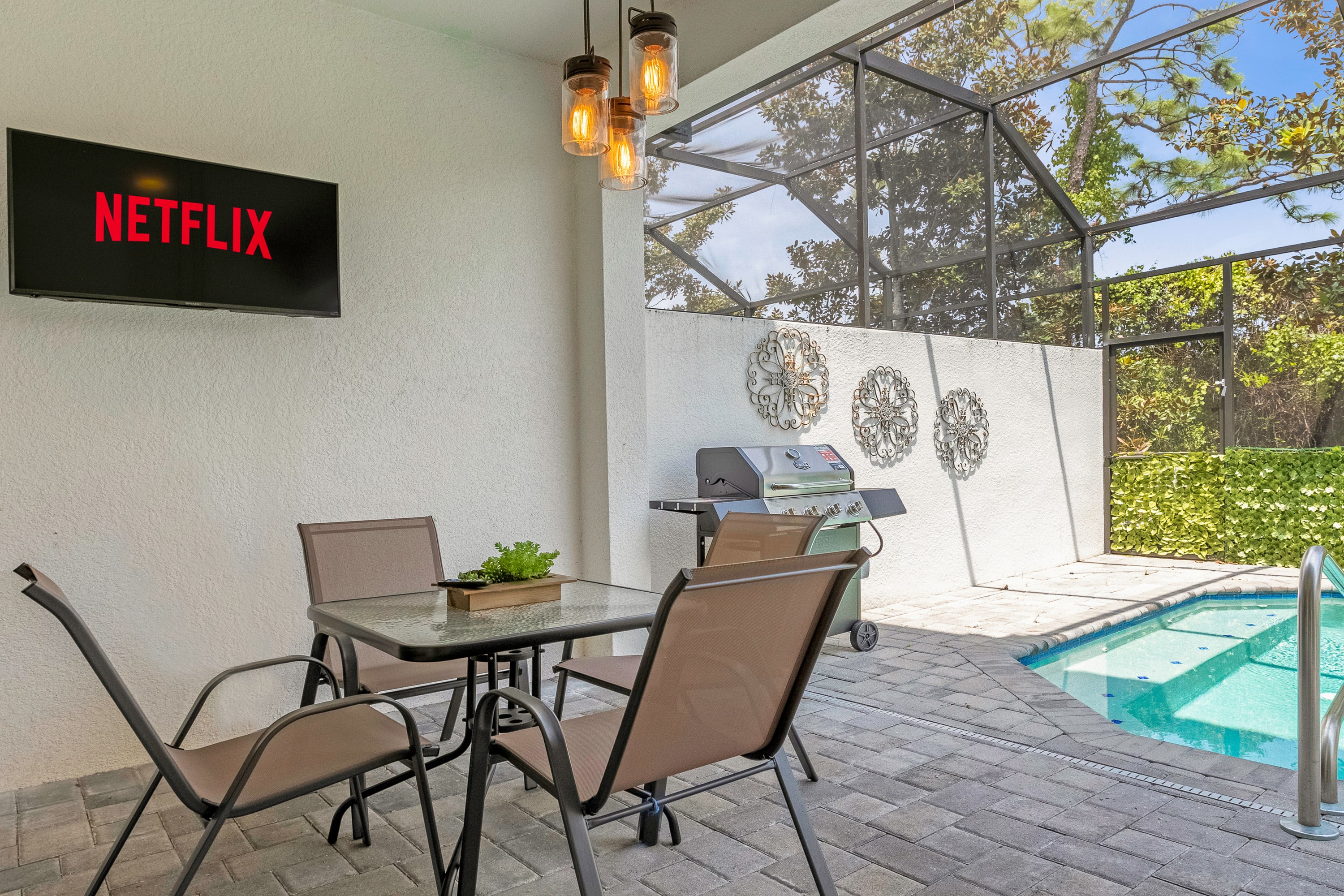 A screened-in patio area features a glass-top dining table with four chairs and a gas BBQ grill, set on light-colored pavers. A wall-mounted TV displays Netflix, and decorative metal wall art adorns the white wall, with the private pool visible to the right.