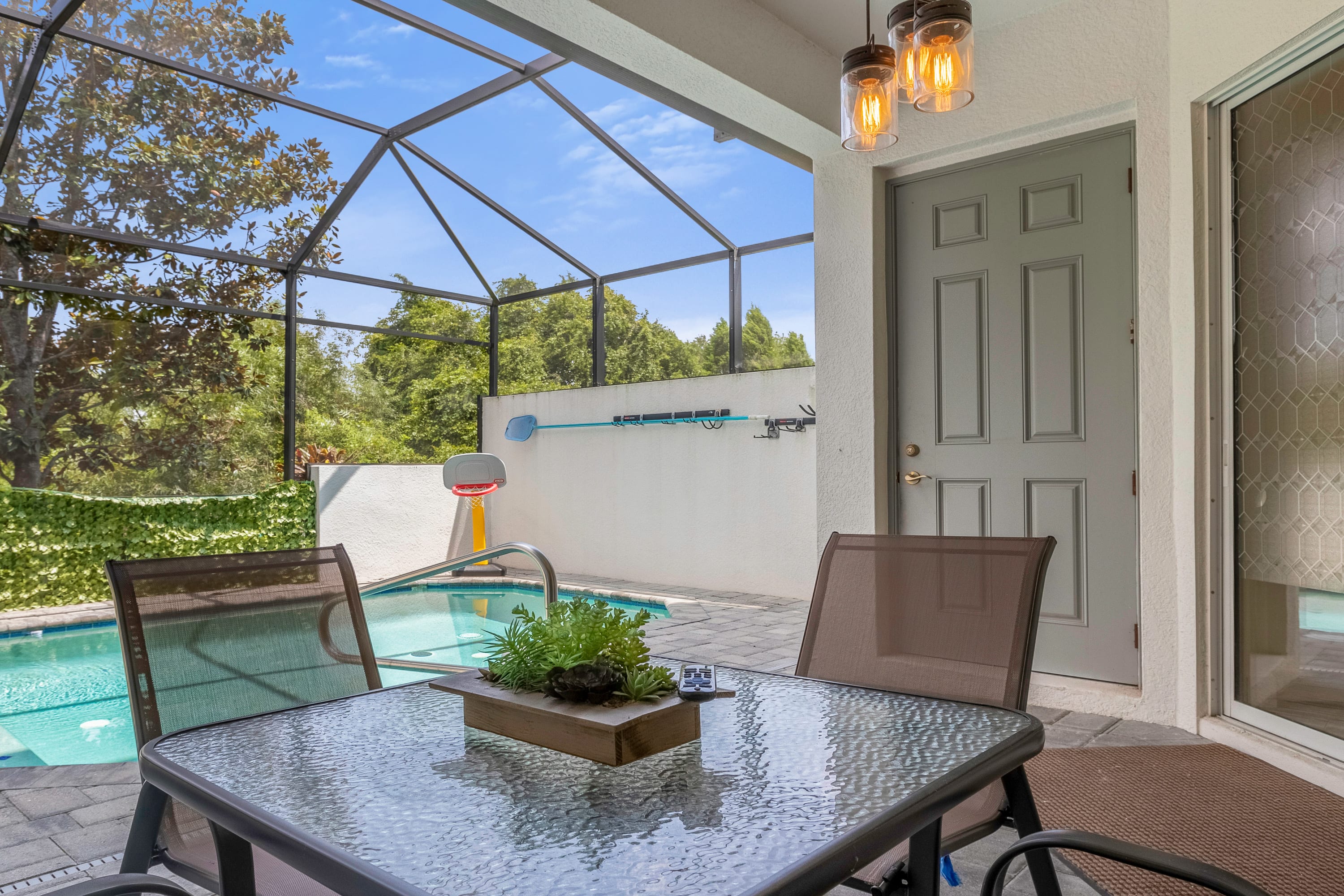 A screened-in patio area features a glass-top dining table with four chairs, overlooking a private pool. The space is enclosed by a dark screen, with a light-colored wall and a gray door leading into the house, and trees visible outside the enclosure.