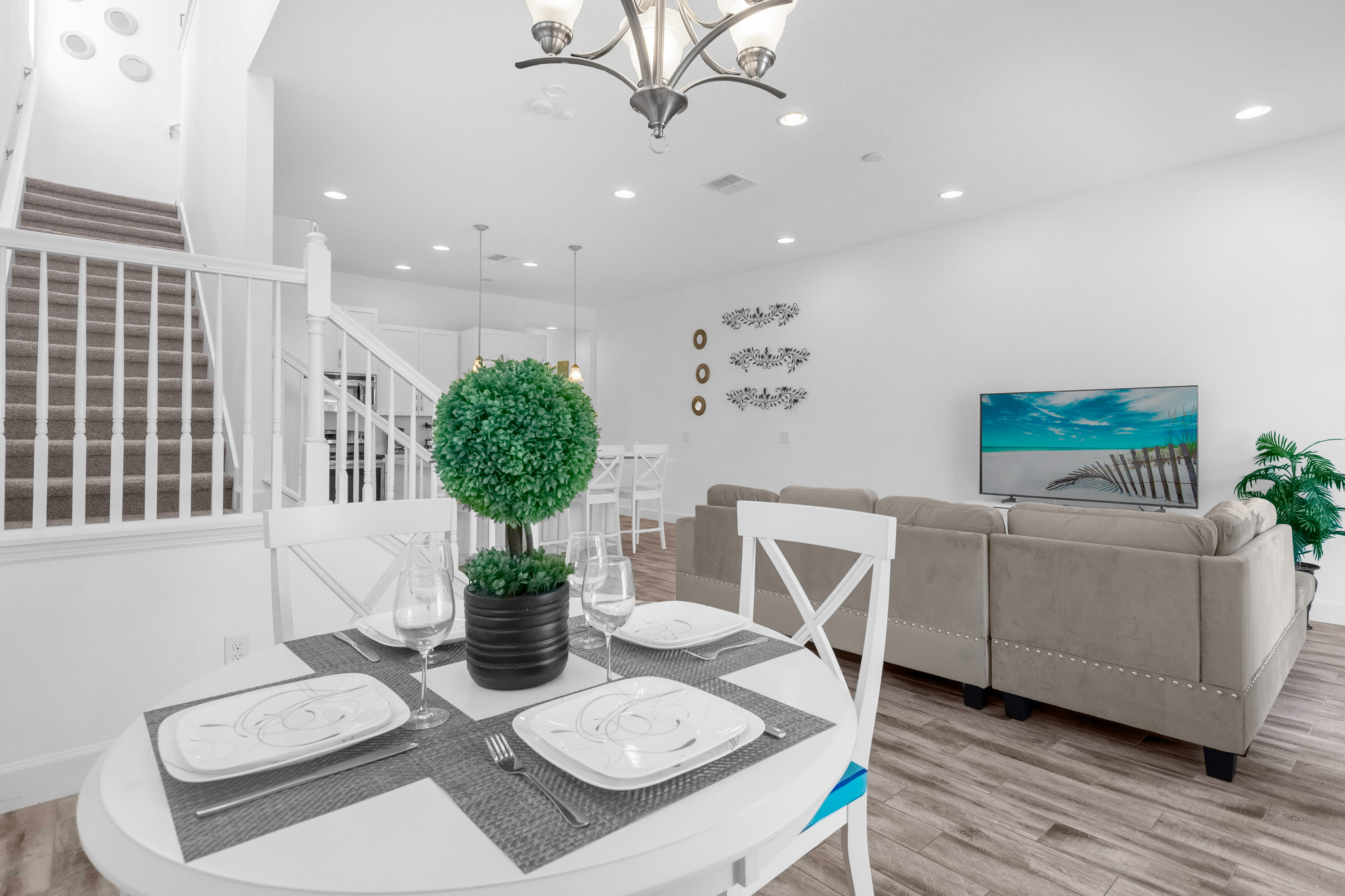 White round dining table with four chairs, set with plates and glasses, featuring a green plant centerpiece. It opens to a living area with a large sofa and TV, and a staircase with carpeted steps.