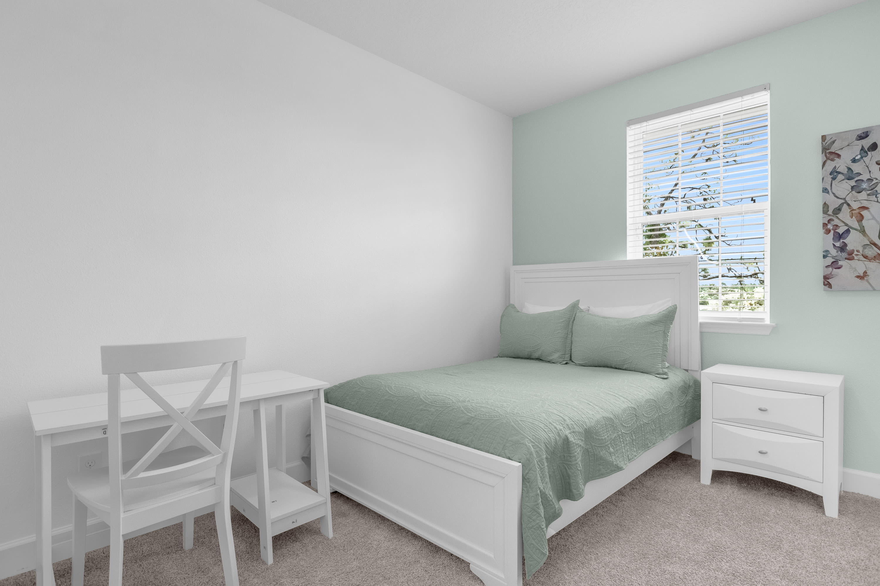 Clean and bright bedroom with white walls and a light green accent wall, featuring a white full-size bed with a matching headboard and light green bedding. A white desk and chair are positioned to the left, and a window with blinds provides natural light.