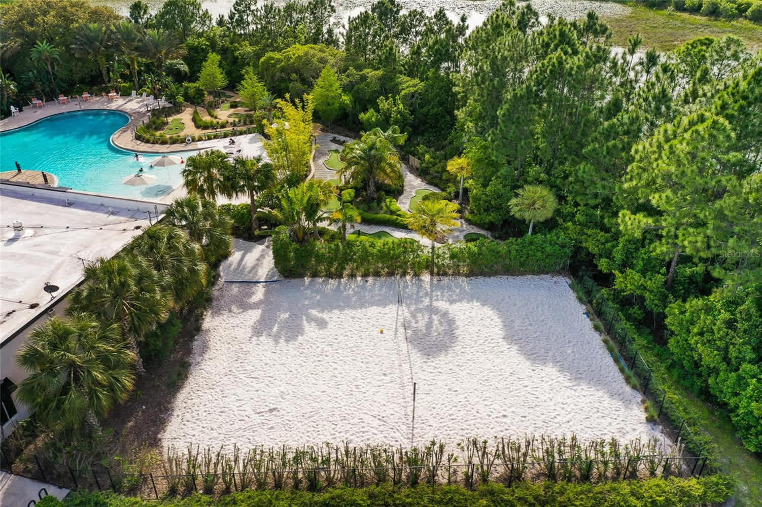 An aerial view of a sandy beach volleyball court, enclosed by a low hedge, with a net in the center. In the background, a large swimming pool and lush green trees are visible, indicating a resort or community recreation area.
