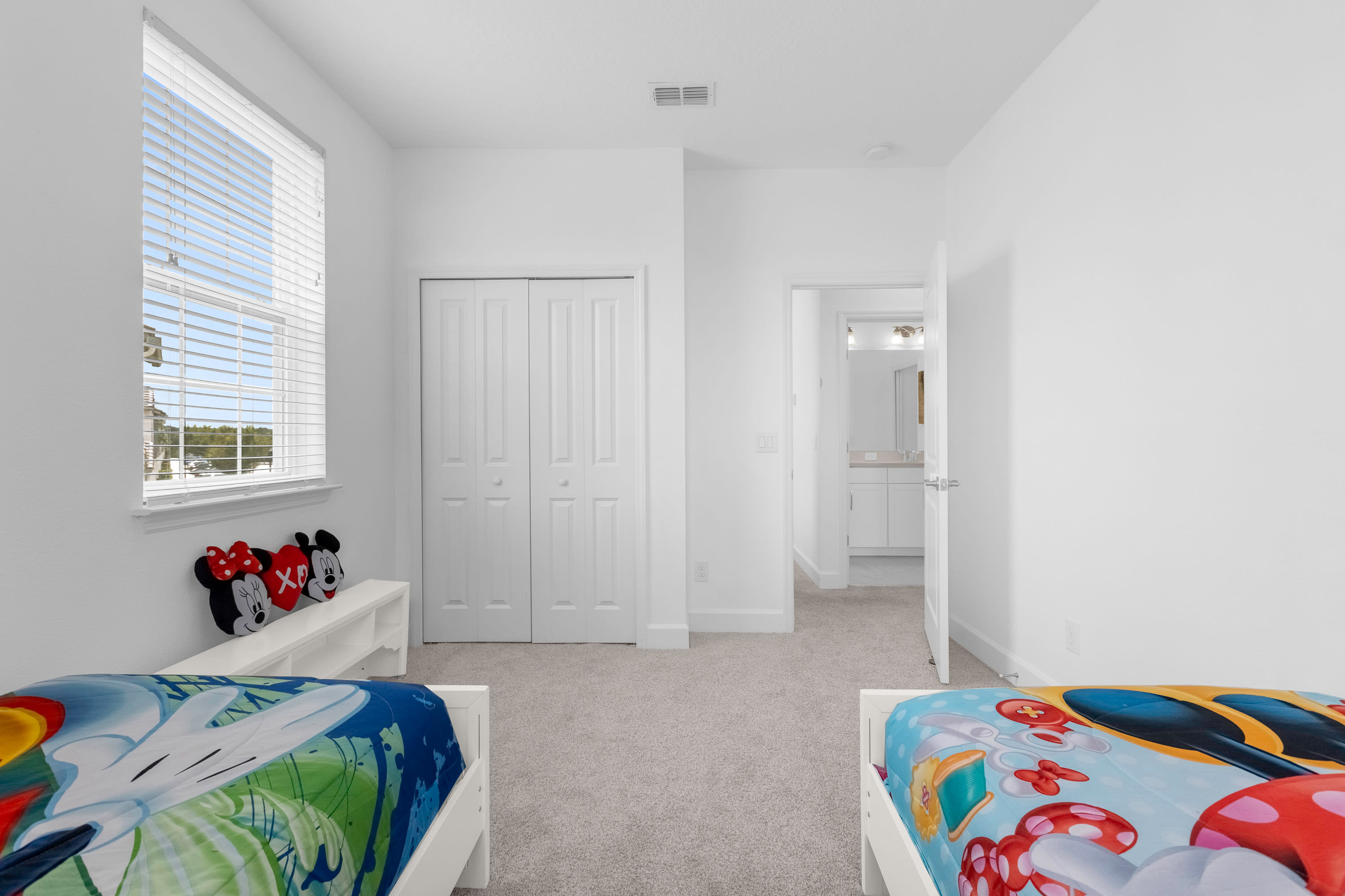 Another angle of the children's bedroom, showing a white bi-fold closet door and a low white shelf with Mickey and Minnie Mouse plush toys. An open doorway leads to an adjacent bathroom, maintaining the room's bright and clean aesthetic.