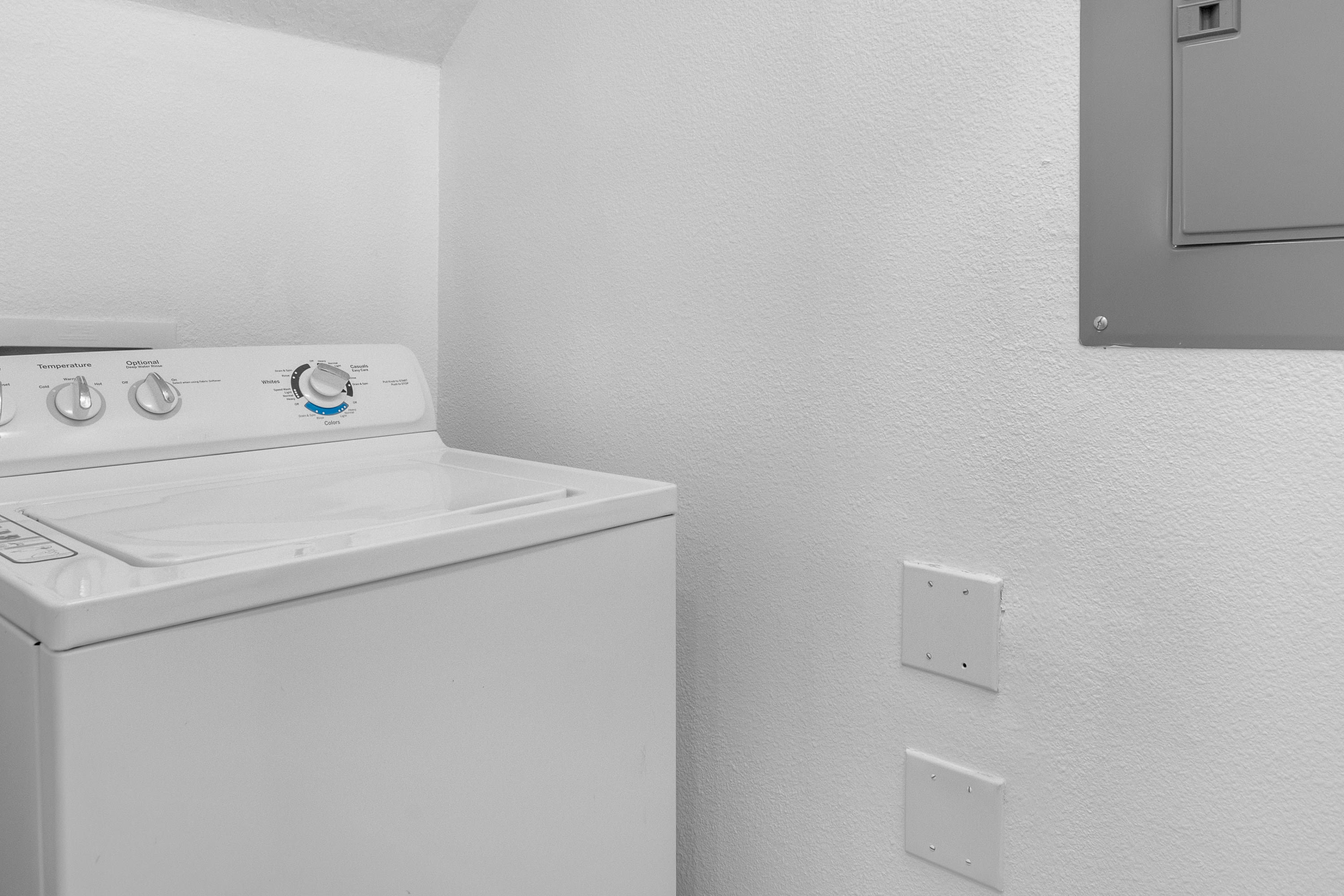 A close-up view of a white top-loading washing machine with control knobs for temperature and water level, set against a white textured wall. Two white electrical outlet covers are visible on the wall below a grey electrical panel.