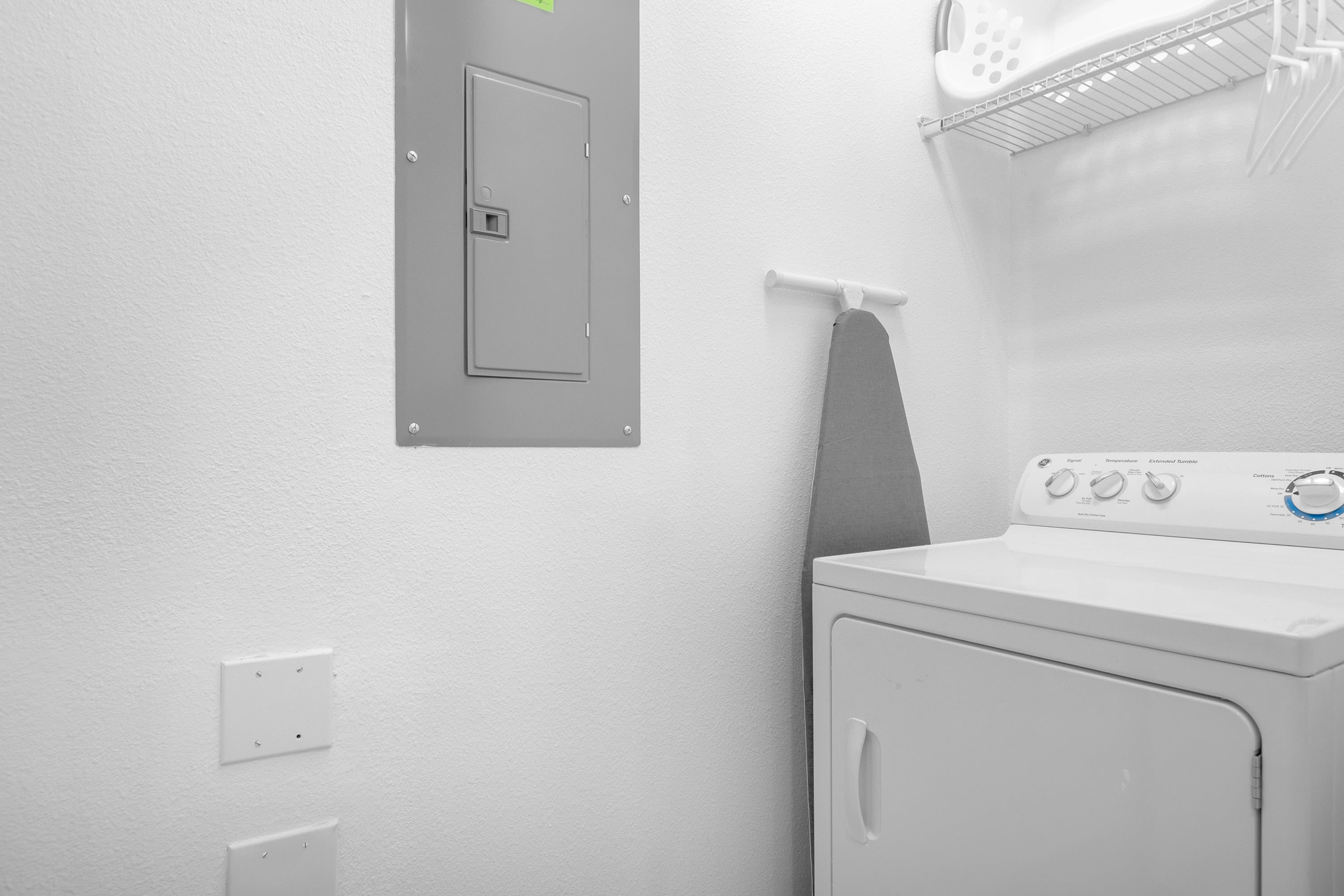 A bright laundry area features a white top-loading washing machine and a dryer, with an ironing board hanging on the wall. A grey electrical panel is mounted on the white textured wall, and a wire shelf with a laundry basket is visible above.