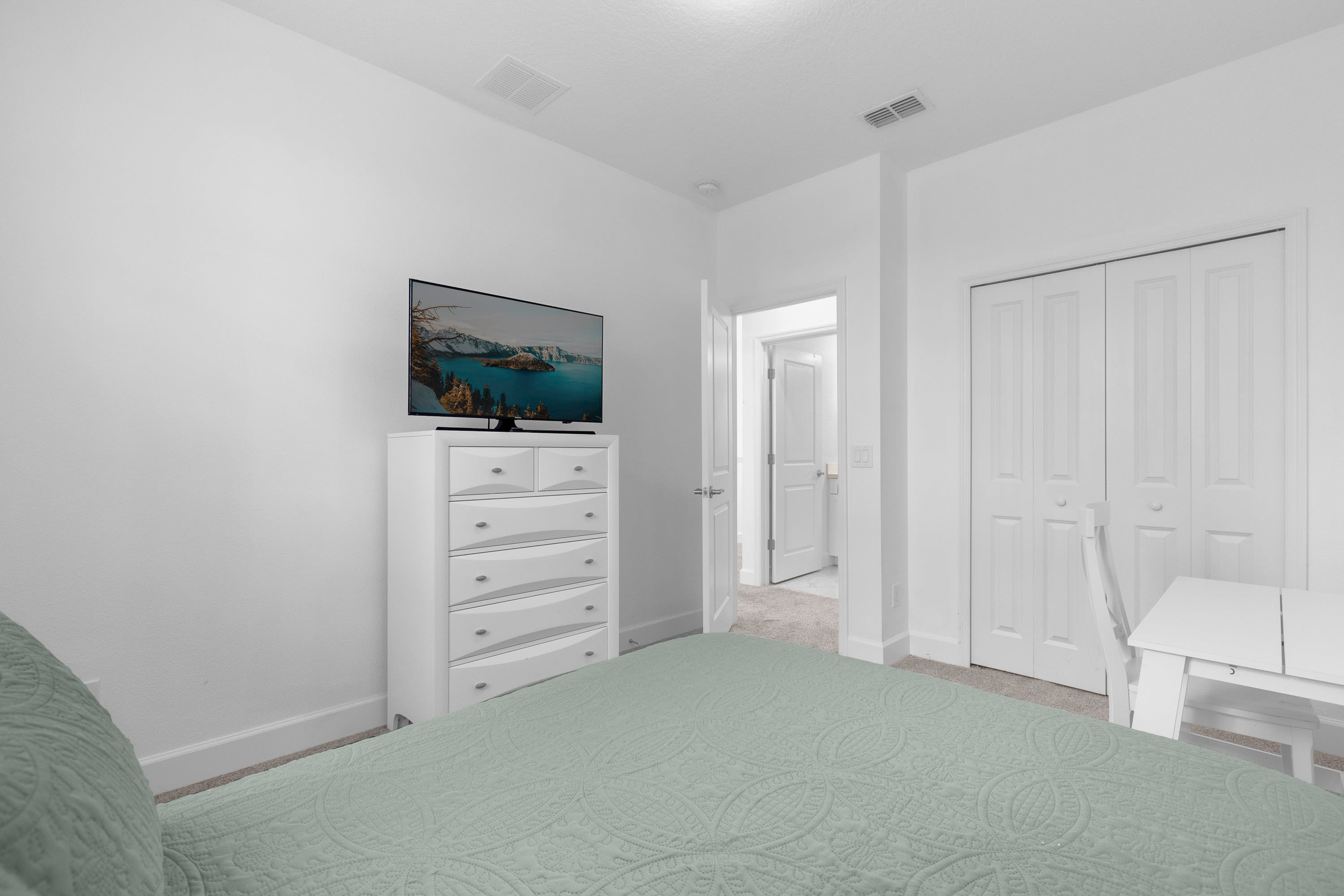 Another view of the bedroom, showcasing a white dresser with multiple drawers and a flat-screen TV mounted on top. A white bi-fold closet door is visible, and the room maintains its clean, minimalist aesthetic with white walls and carpet.