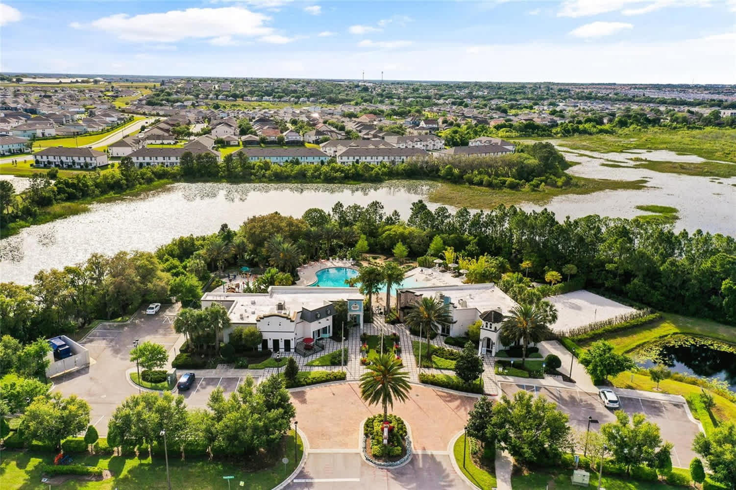 An expansive aerial view of a resort community featuring a large central clubhouse area with multiple pools, surrounded by lush green landscaping and a lake. Residential buildings and a vast natural landscape are visible in the background under a partly cloudy sky.