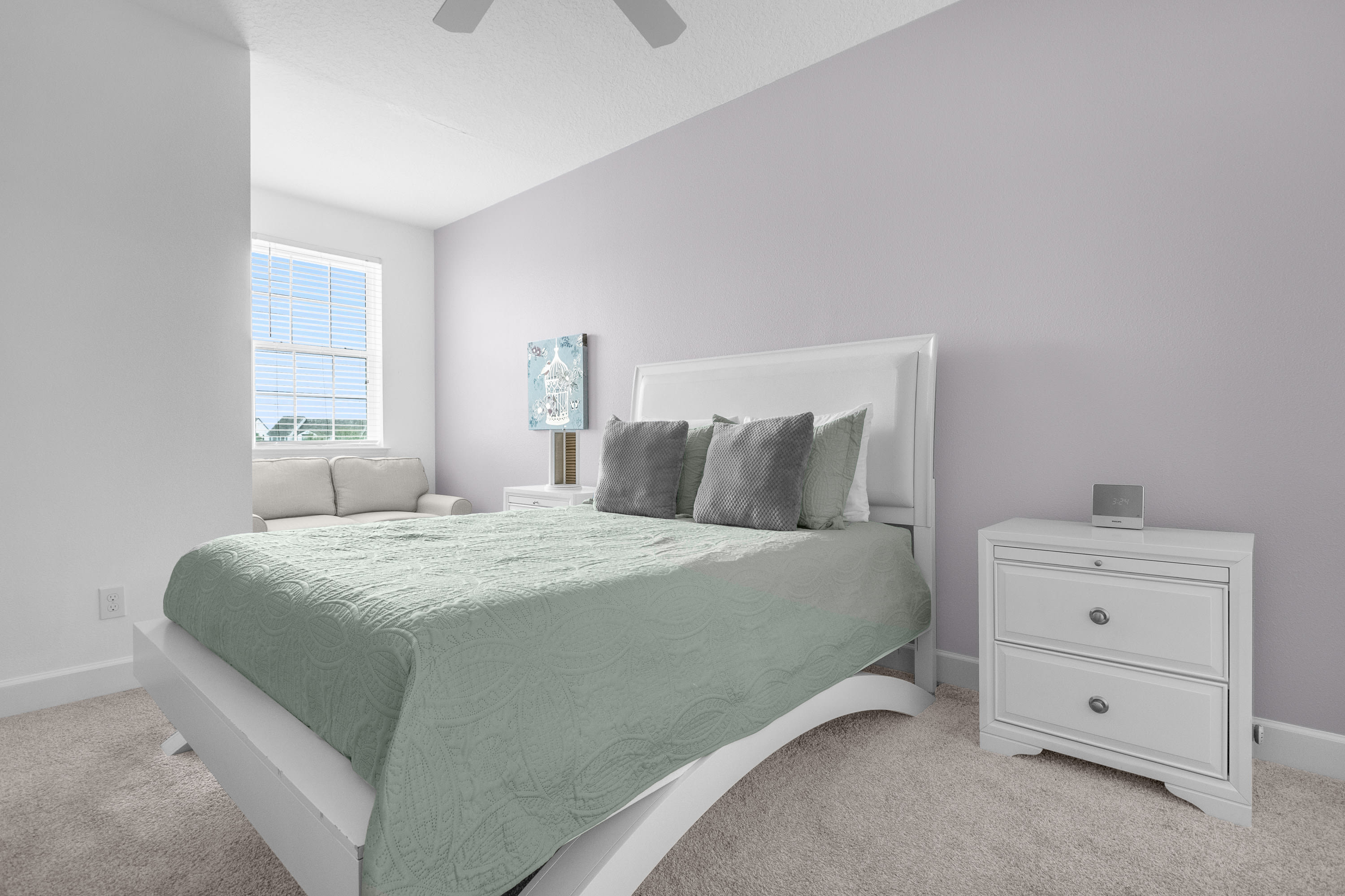 Serene bedroom with light gray walls and plush carpet, featuring a white sleigh-style queen bed with a light green comforter and gray pillows. A white nightstand with drawers sits beside the bed, and a window with blinds offers natural light.