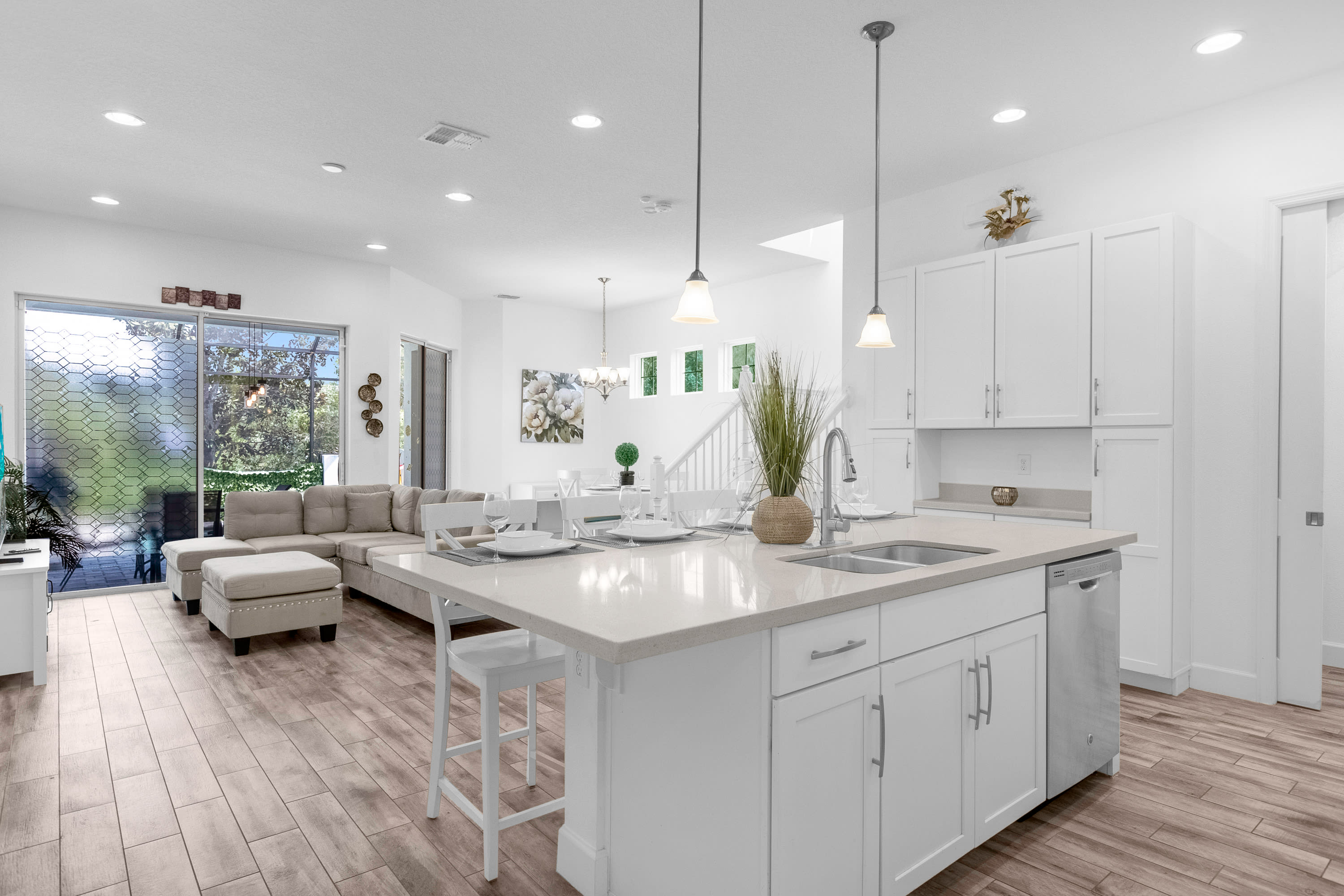 Modern white kitchen with a large island featuring a sink, dishwasher, and seating for four. The open-concept space connects to a living area with a sofa and a dining area with a table and chairs.