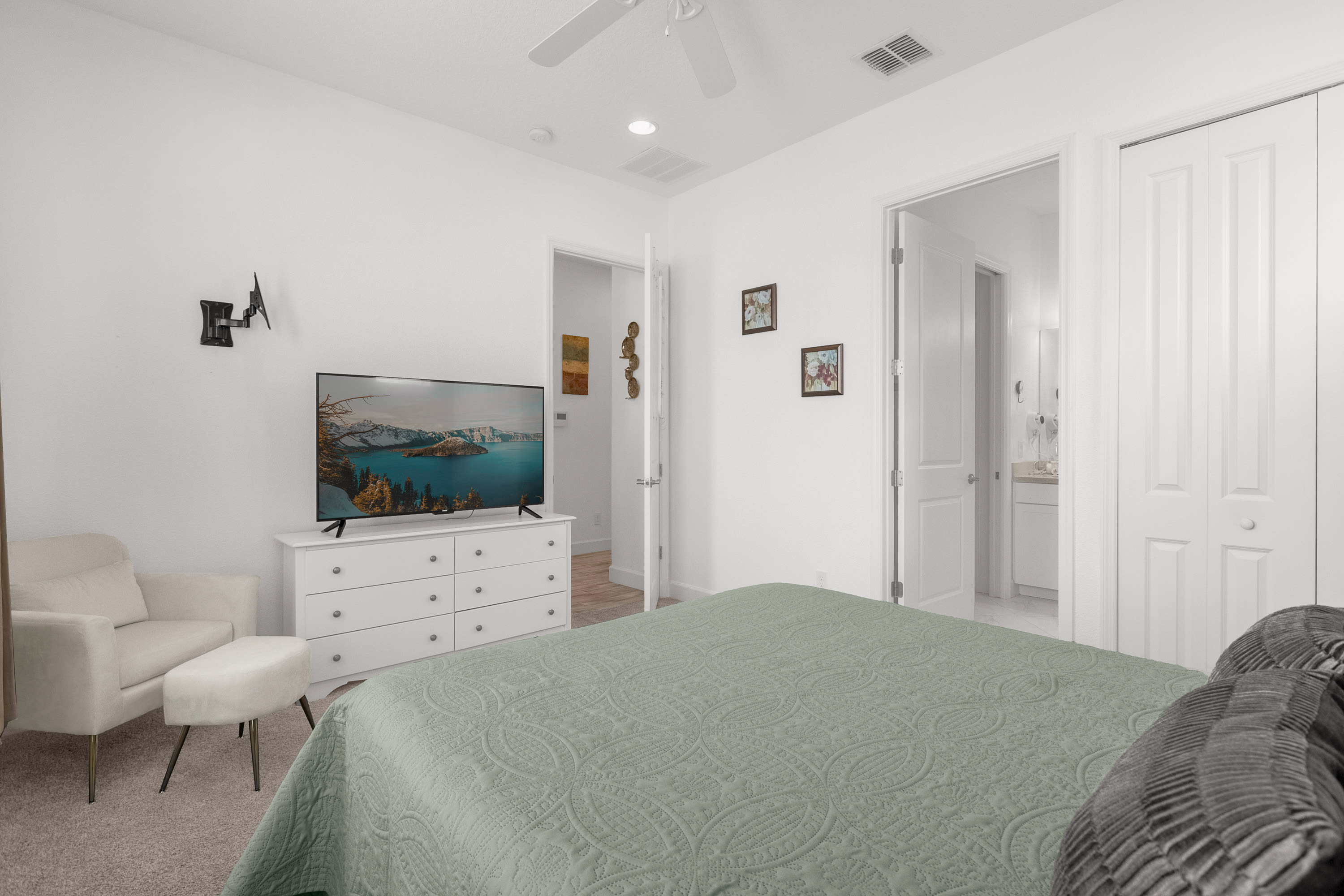 View of bedroom 2 showing a large flat-screen TV on a white dresser, an armchair with an ottoman, and a glimpse into an adjacent bathroom. The room has white walls and a carpeted floor.