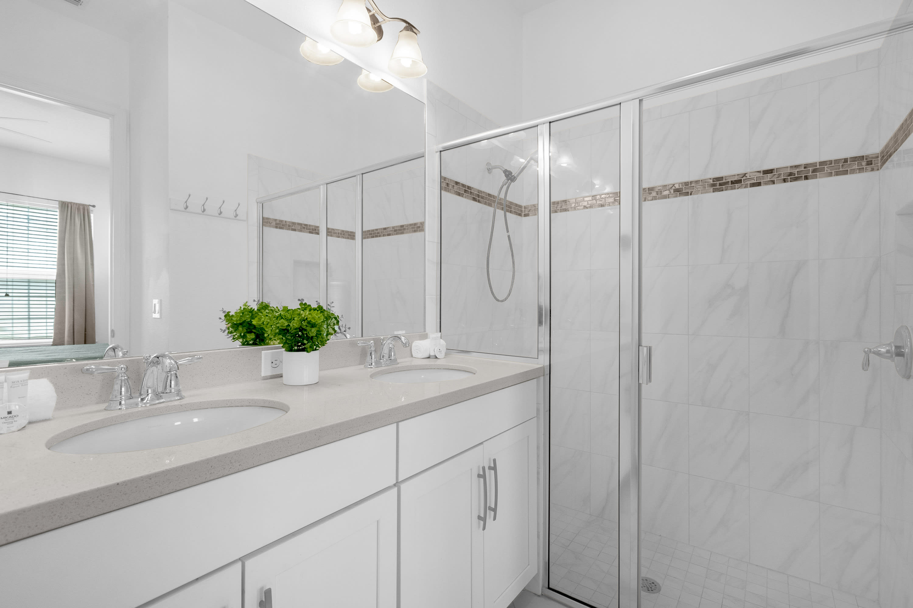 Bright white bathroom featuring a double vanity with light quartz countertops, chrome fixtures, and a large mirror. A spacious walk-in shower with white subway tiles and a decorative mosaic band is visible on the right.