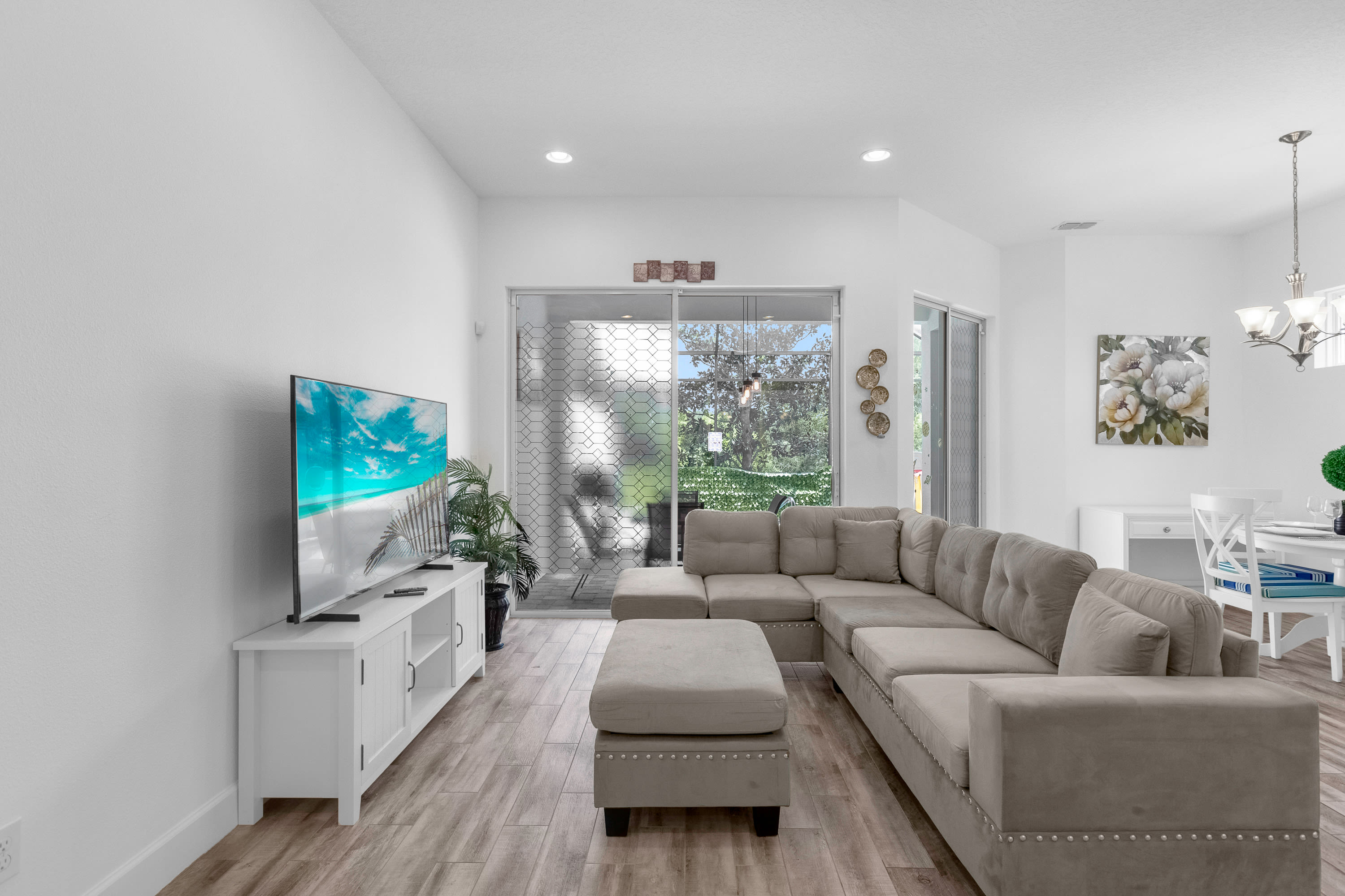 A spacious living room features a large L-shaped sectional sofa and ottoman in a neutral tone, facing a wall-mounted flat-screen TV on a white console. Wood-look tile flooring extends throughout the open space, with a large sliding glass door providing access to the outdoor area.