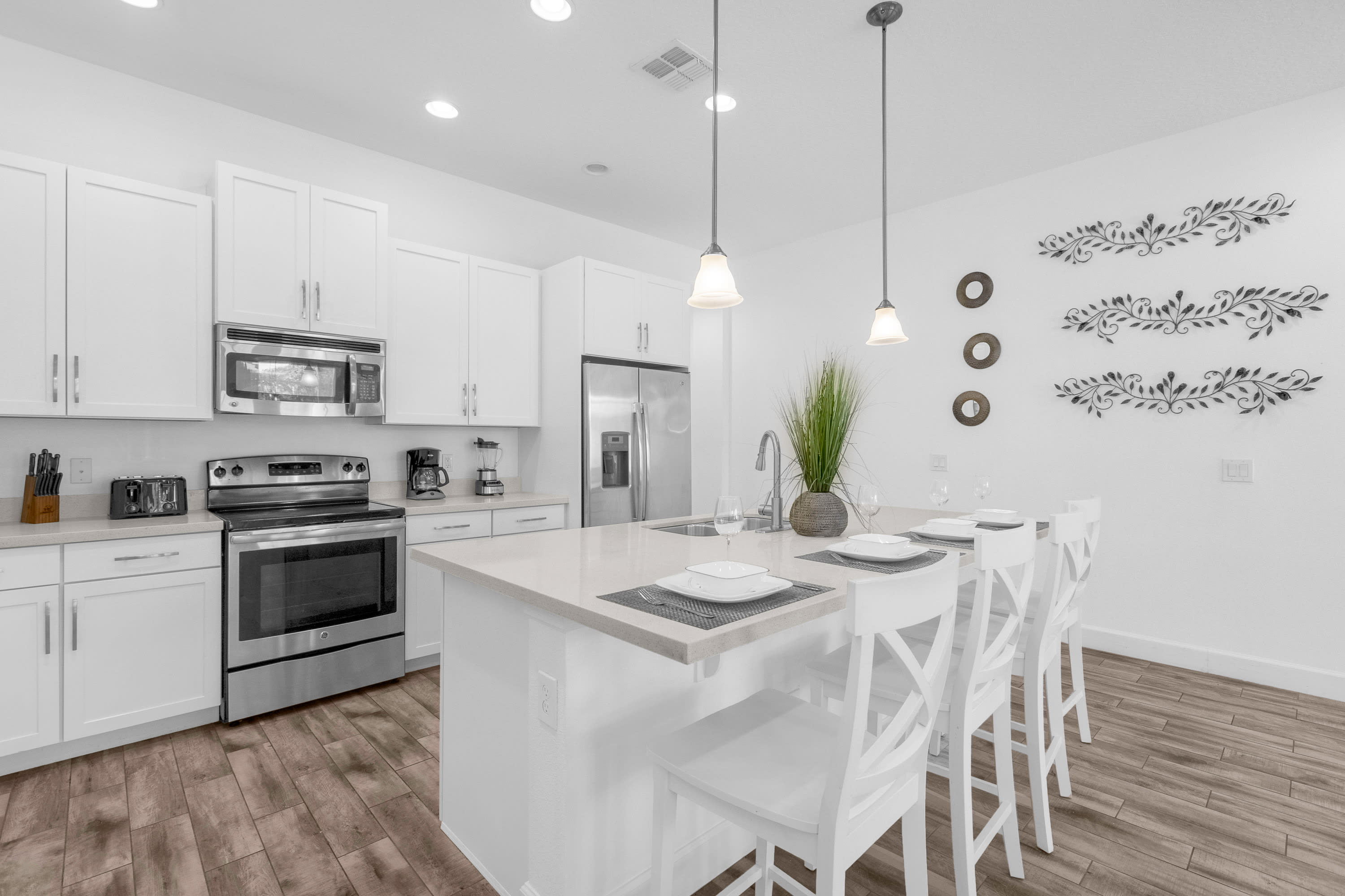 Contemporary white kitchen with a large island featuring a sink and seating for four with white cross-back stools. White shaker cabinets and stainless steel appliances are visible in the background, with decorative wall art.