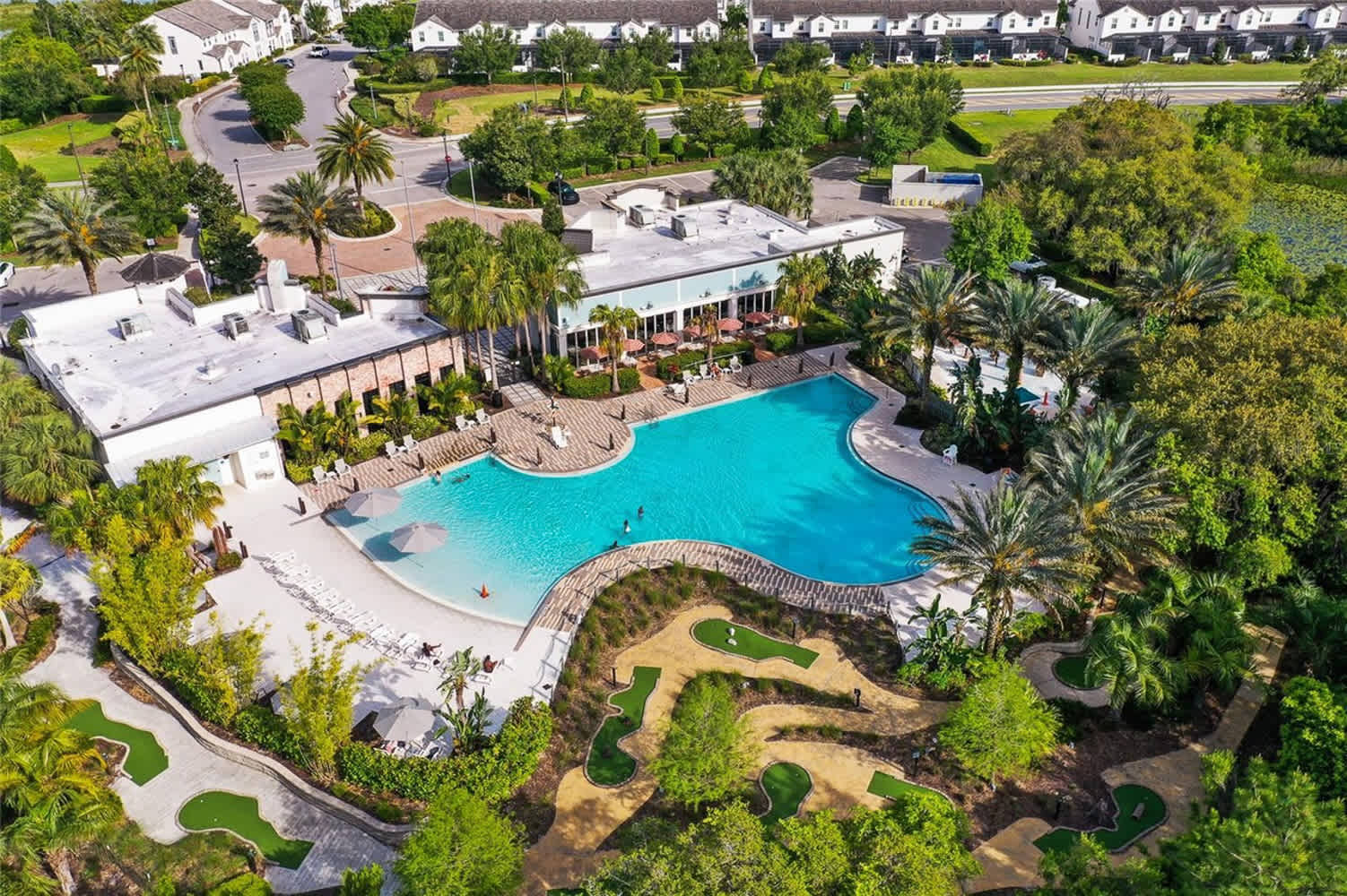 An aerial view showcases a large, irregularly shaped communal swimming pool surrounded by lounge chairs and umbrellas, set within a resort-style complex. Lush green landscaping, palm trees, and buildings with white roofs are visible, along with a miniature golf course.