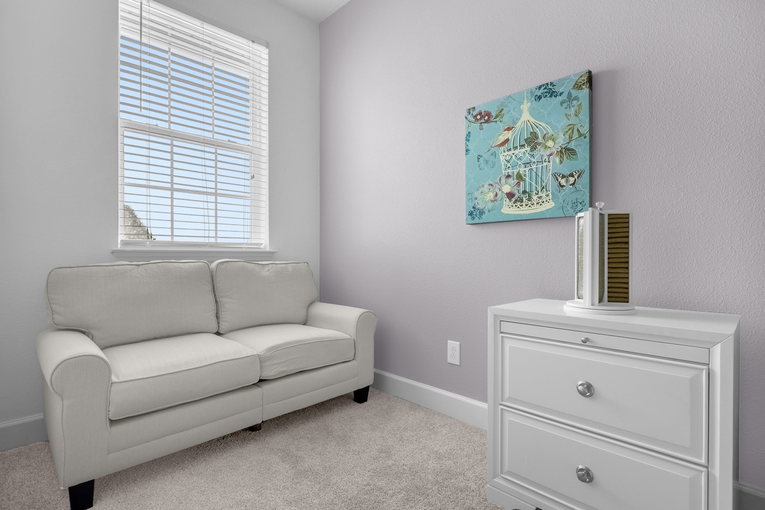 Cozy sitting area with light gray walls and carpet, featuring a cream-colored two-seater sofa with dark legs positioned under a window with blinds. A white dresser with drawers and a decorative birdcage artwork hangs on the wall.