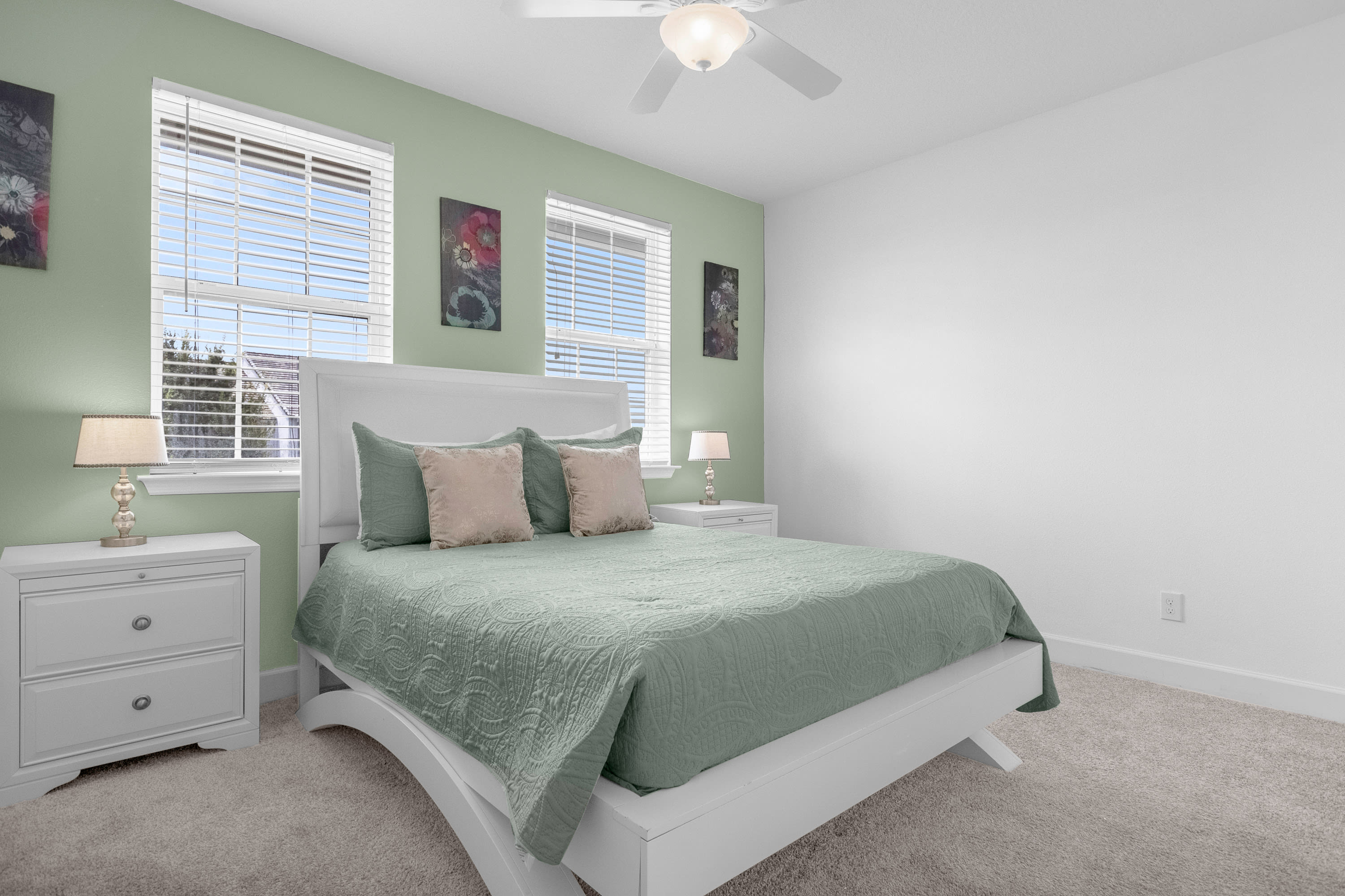 Bright bedroom with a light green accent wall and white walls, featuring a white queen-sized bed with a green comforter and two nightstands with lamps. Two windows with blinds provide natural light.