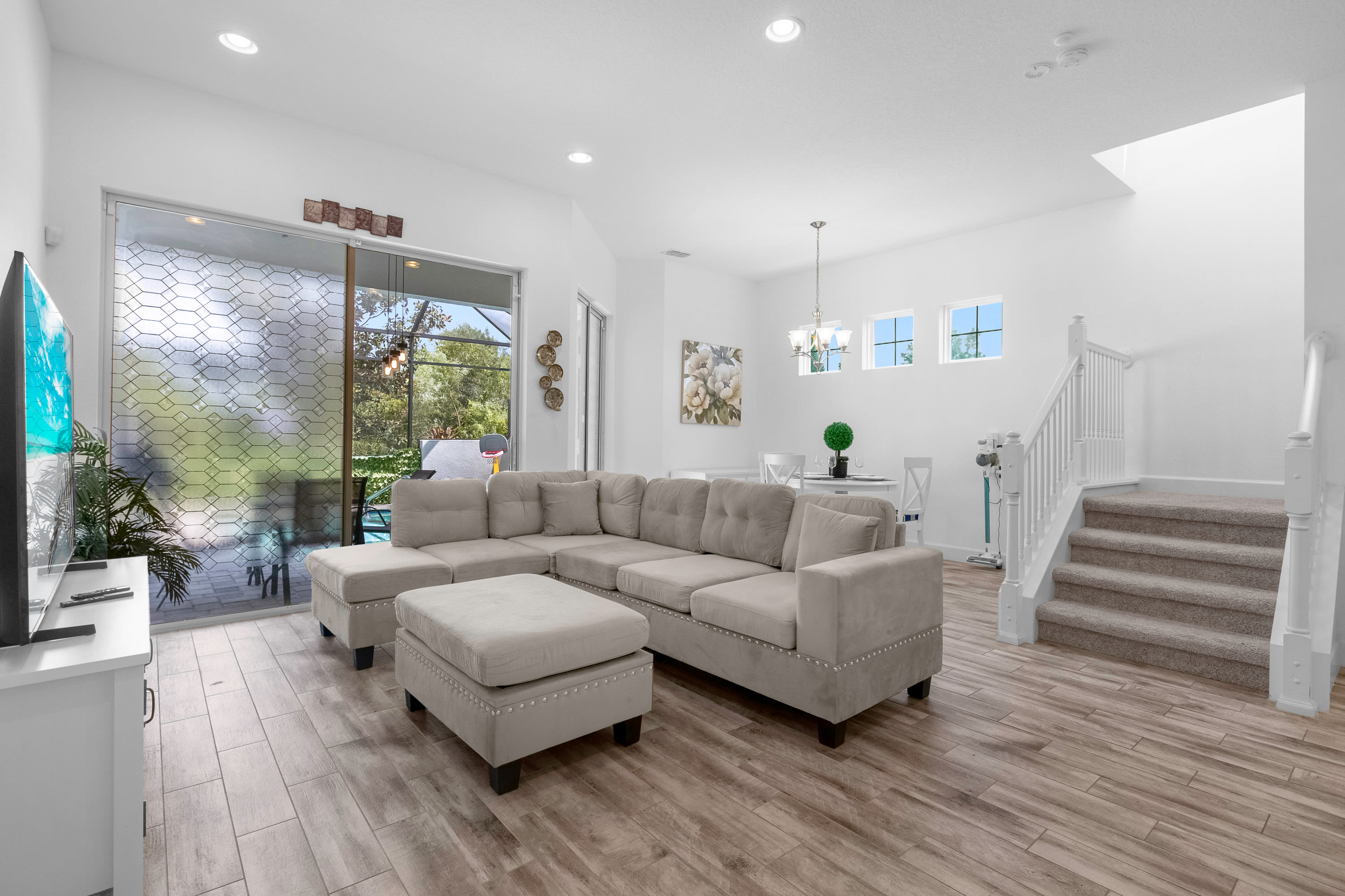 An open-concept living space features a large L-shaped sectional sofa and ottoman in a neutral tone, set on wood-look tile flooring. A large sliding glass door leads to an outdoor area, and a dining table with chairs is visible in the background, next to a carpeted staircase.