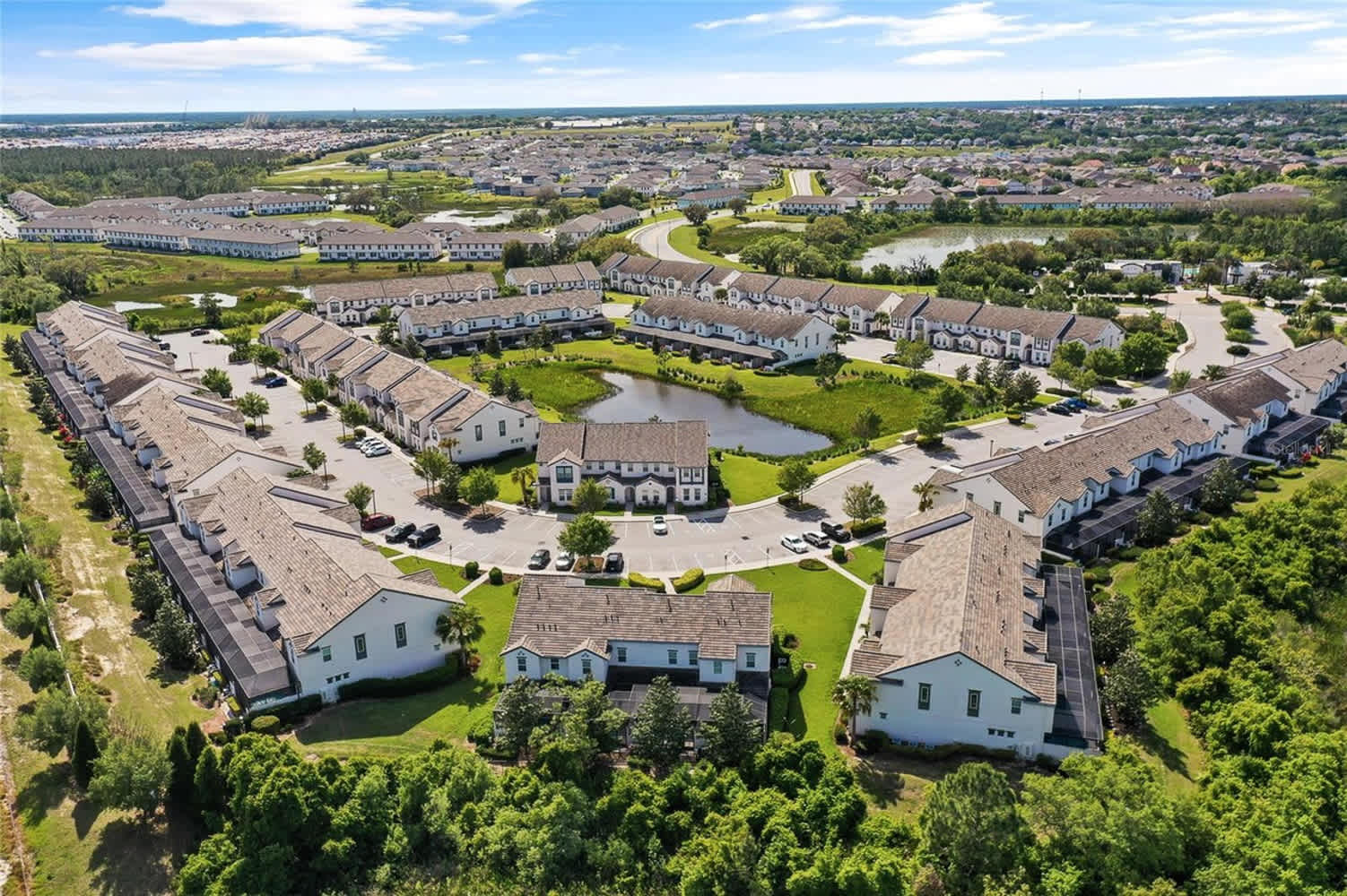 An aerial view of a residential community with rows of modern townhouses and single-family homes, arranged around a central pond and green spaces. The houses feature light-colored exteriors and dark roofs, surrounded by well-maintained landscaping.