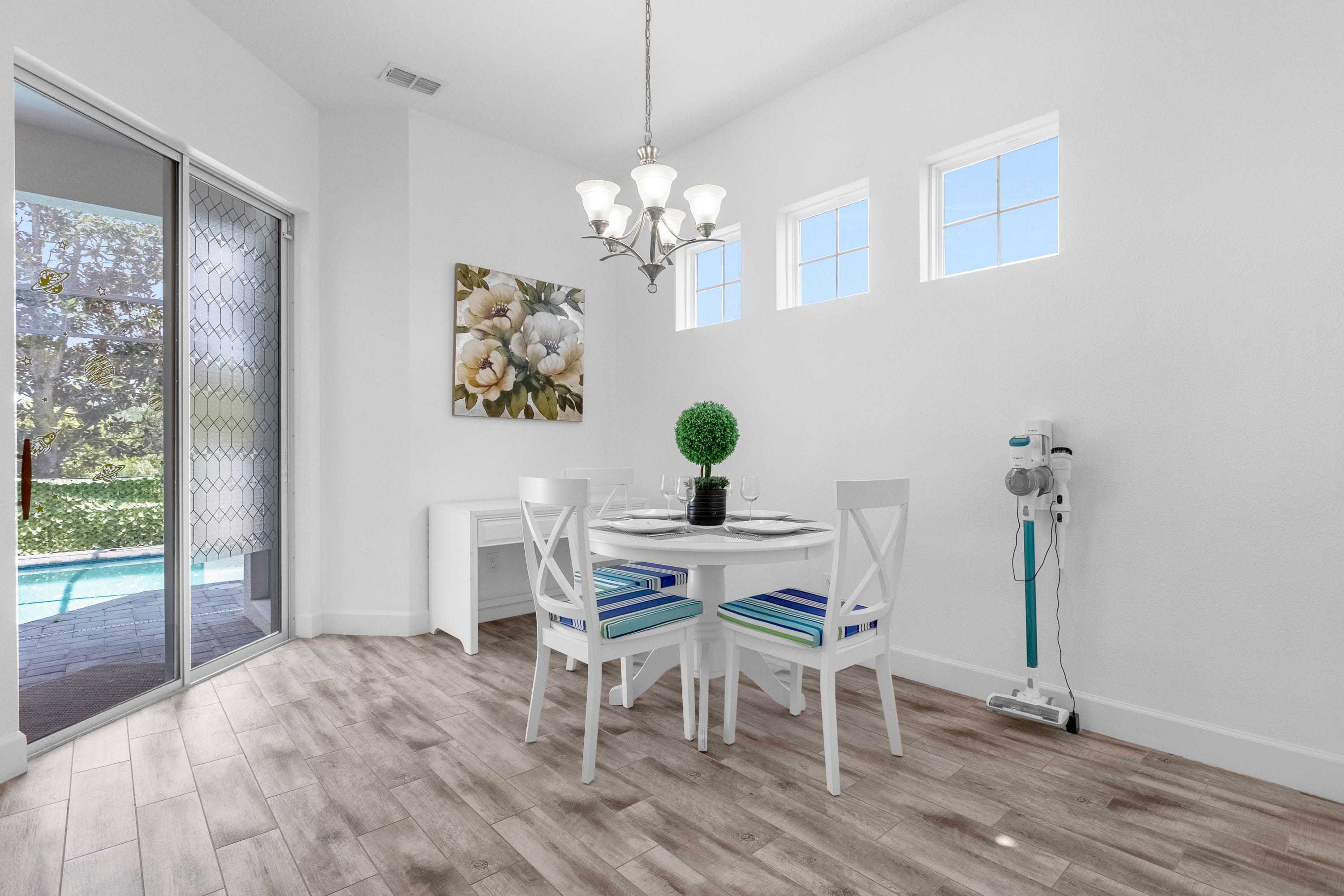 Bright dining area with a white round table and four chairs with blue and white striped cushions, set under a chandelier. A sliding glass door leads to an outdoor pool area, and three high windows let in natural light.