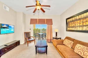 A bright living room features a brown sofa with decorative pillows, a dark wood coffee table, and a wall-mounted TV. A sliding glass door leads to a balcony with outdoor seating, under a ceiling fan.