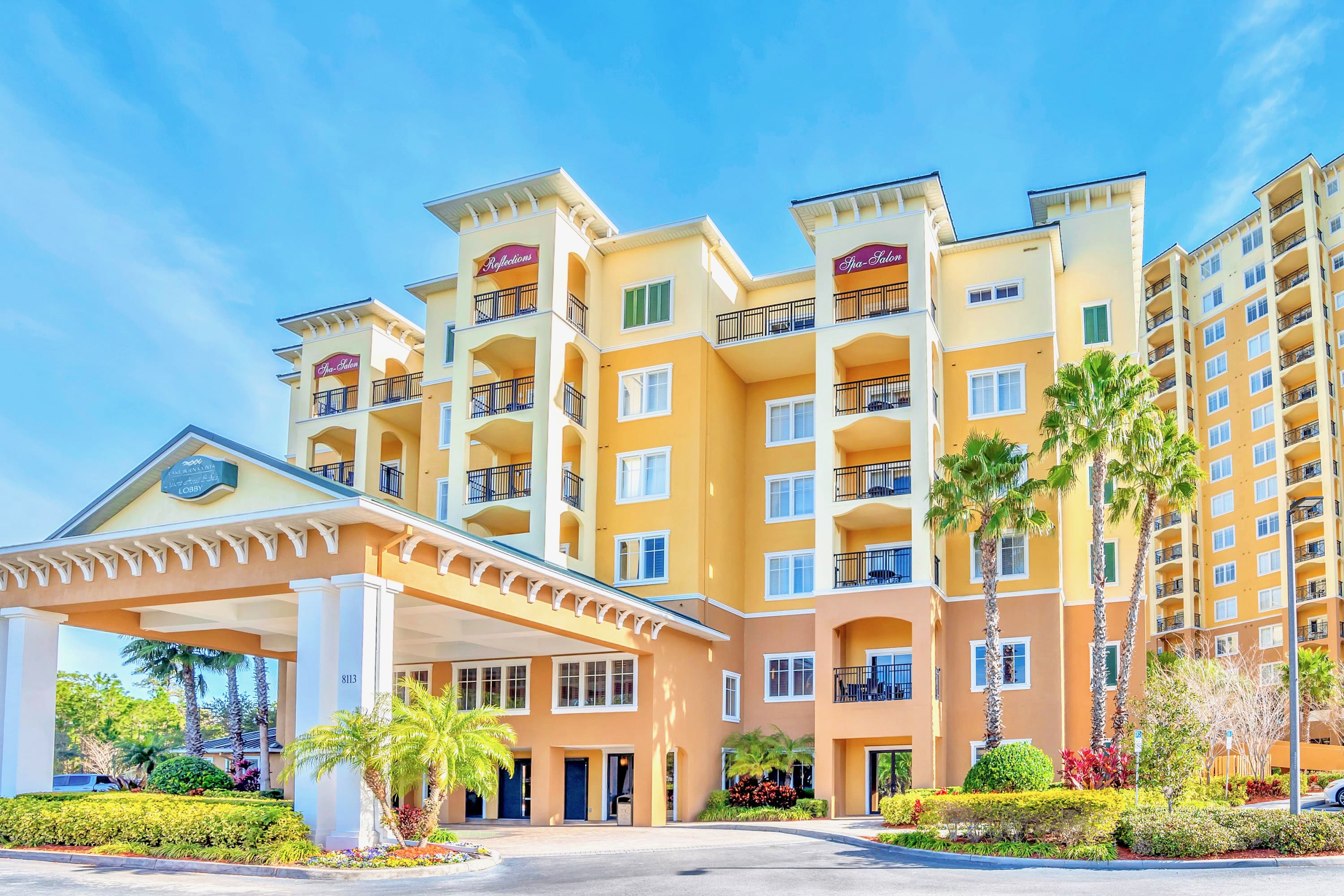 The grand entrance of a multi-story resort building features a large covered drive-through portico with columns and intricate architectural details. The building is painted in warm yellow and orange tones, surrounded by palm trees and lush landscaping.