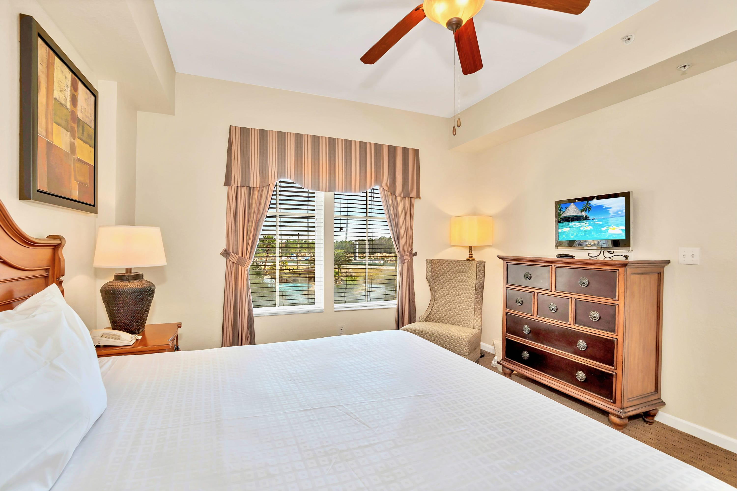 Master bedroom featuring a king-sized bed, a dark wood dresser with a television, a patterned armchair, and a window with blinds and striped curtains overlooking a waterfront.