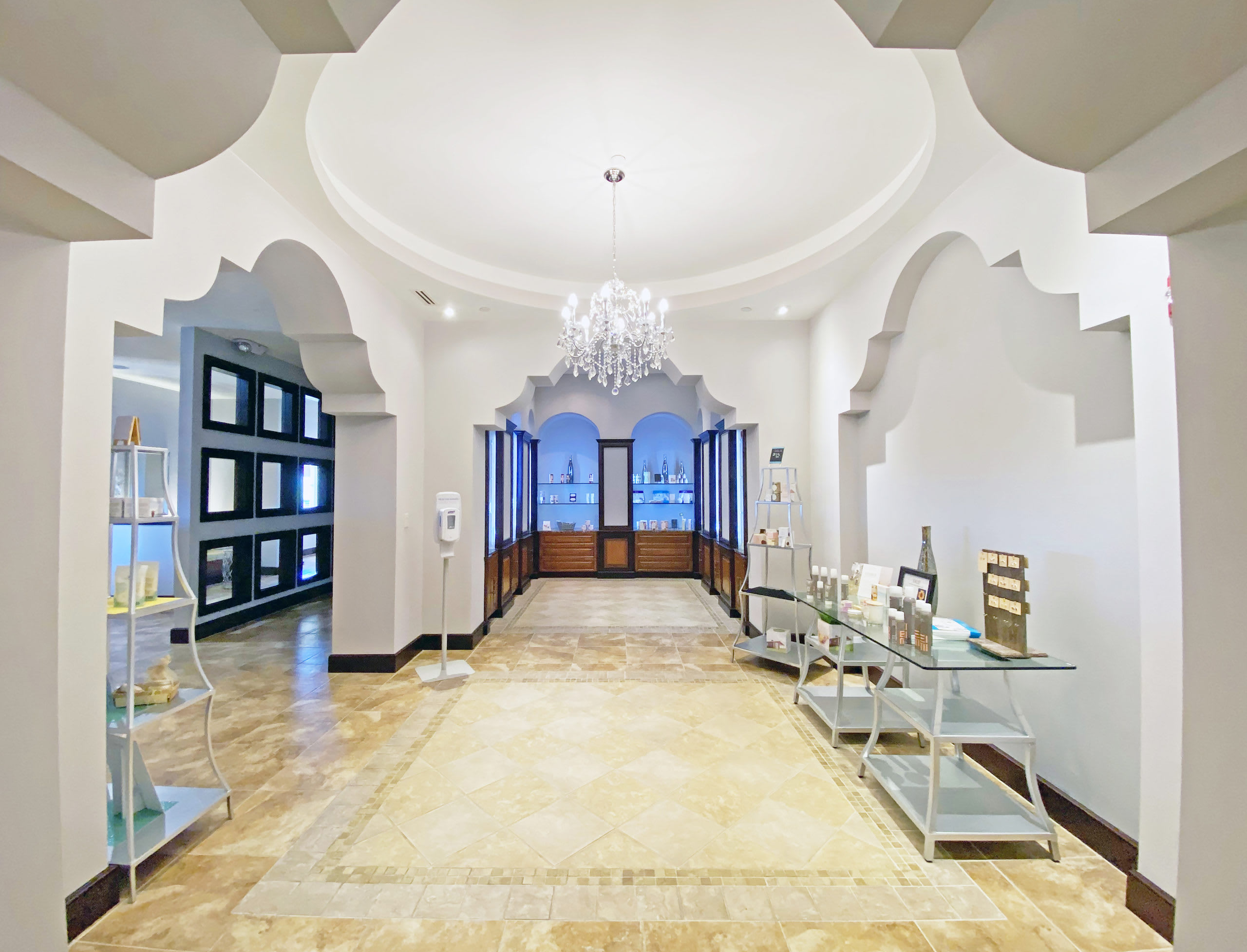 A luxurious interior hallway features ornate arched doorways and a vaulted ceiling with a sparkling chandelier, leading to display cases filled with beauty products. The floor is tiled with a decorative pattern, and mirrors line one wall.