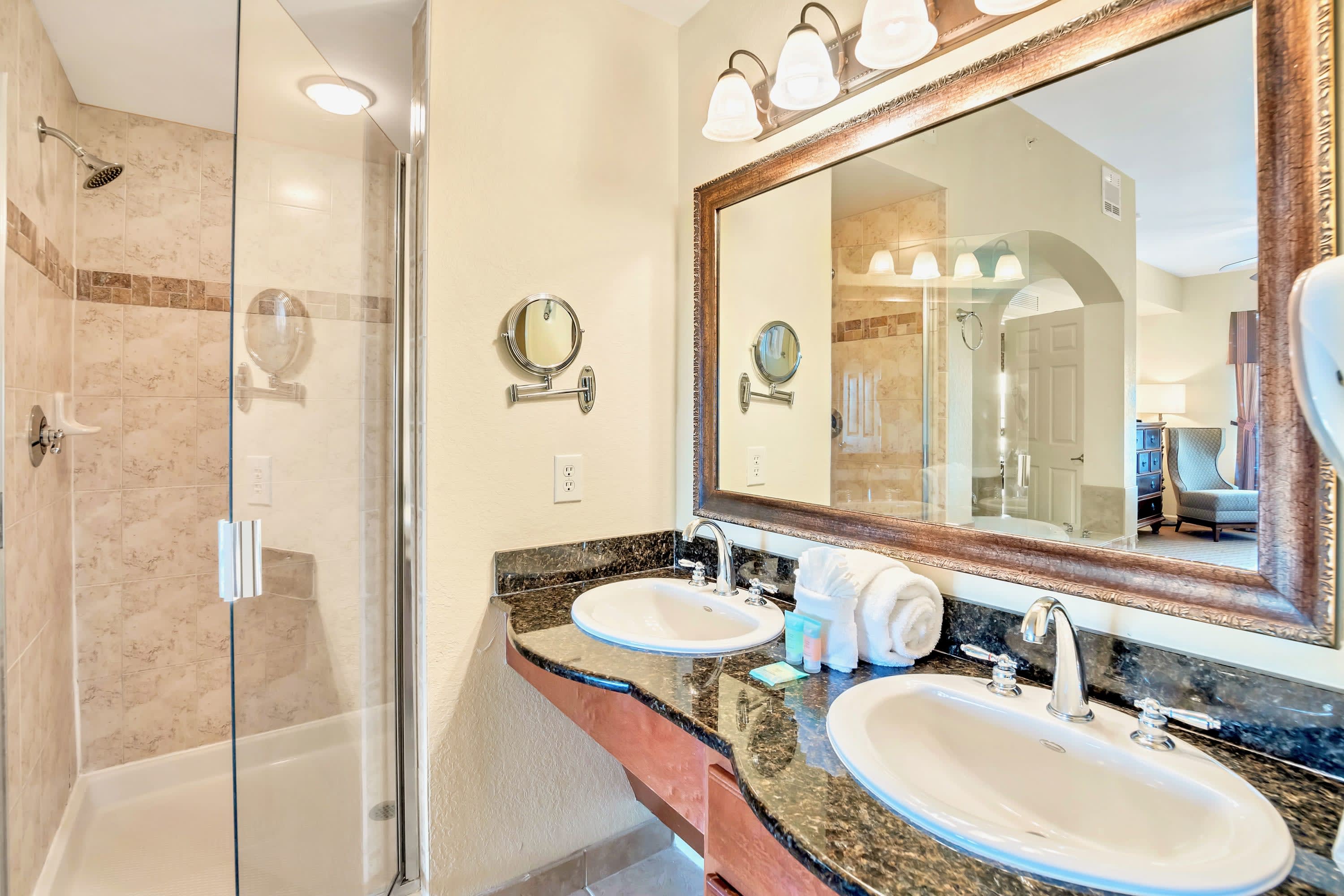 Close-up of the double vanity in the en-suite bathroom, featuring two sinks, dark granite countertops, a large framed mirror, and a glass-enclosed shower to the left.
