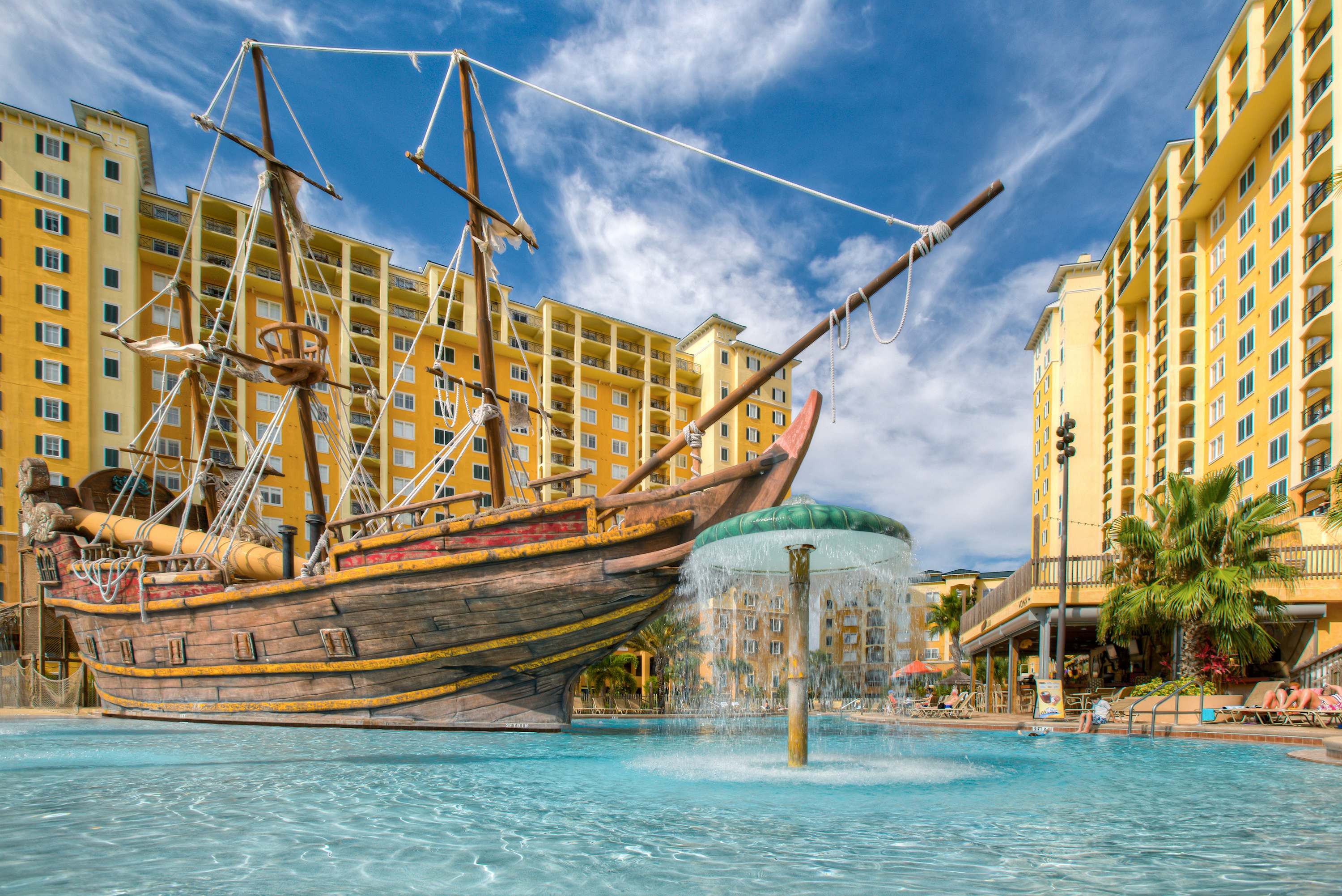 A vibrant swimming pool features a large, detailed wooden pirate ship structure with water features, set against a backdrop of multi-story resort buildings. The pool water is clear and blue under a bright sky.