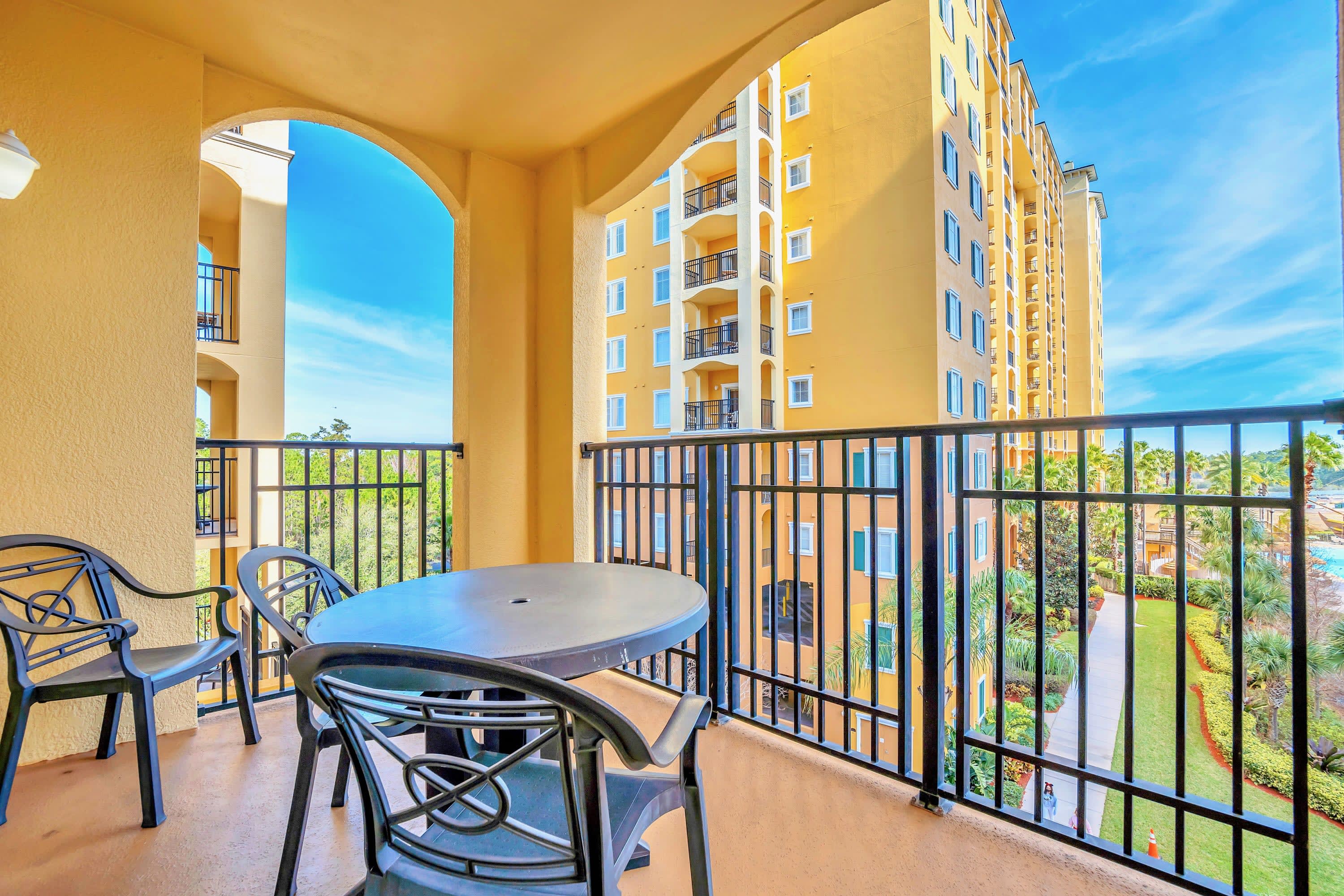 A private balcony with black metal railings offers outdoor seating with a round table and two chairs, providing a view of the resort's yellow buildings and lush landscaping below.