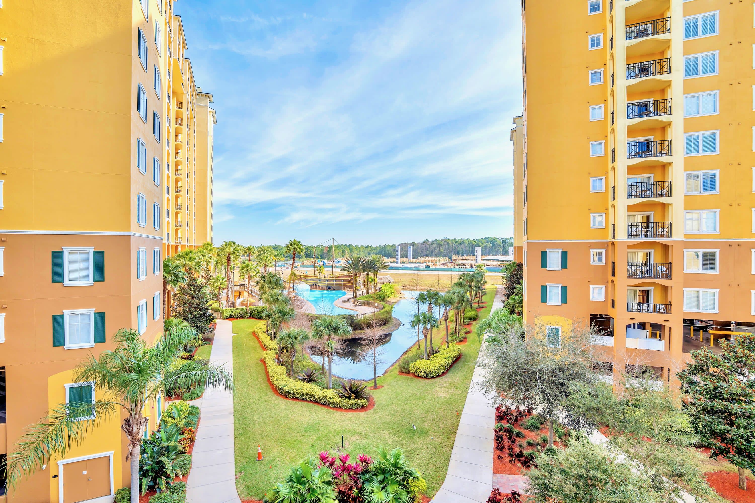 An aerial view showcases a lush resort courtyard with winding pathways, manicured lawns, and tropical palm trees, flanked by two multi-story yellow buildings with balconies. A large swimming pool area is visible in the distance.