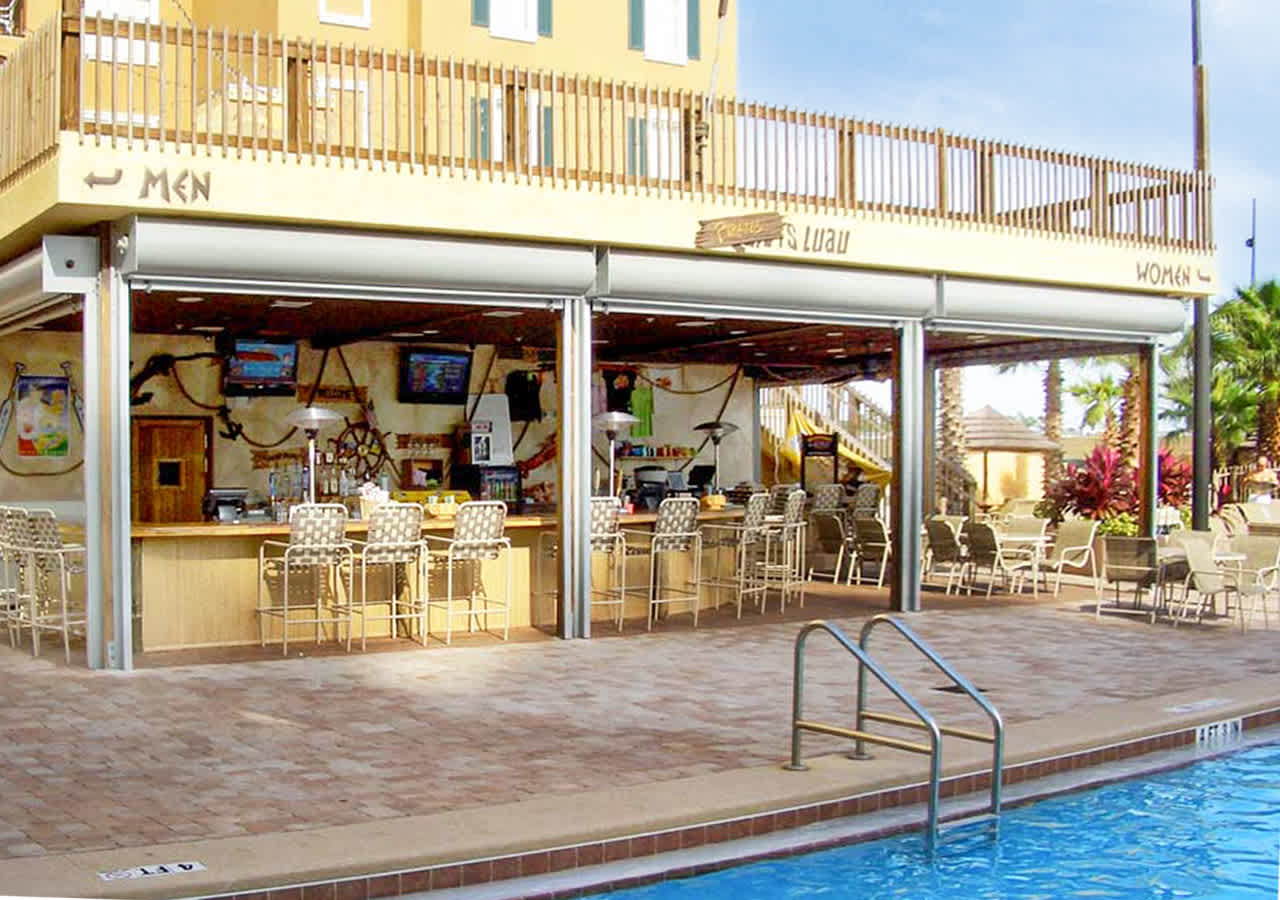A vibrant poolside bar with an open-air design features multiple bar stools and tables, offering a lively spot for guests to relax. The bar is situated next to a swimming pool, with resort buildings visible in the background.