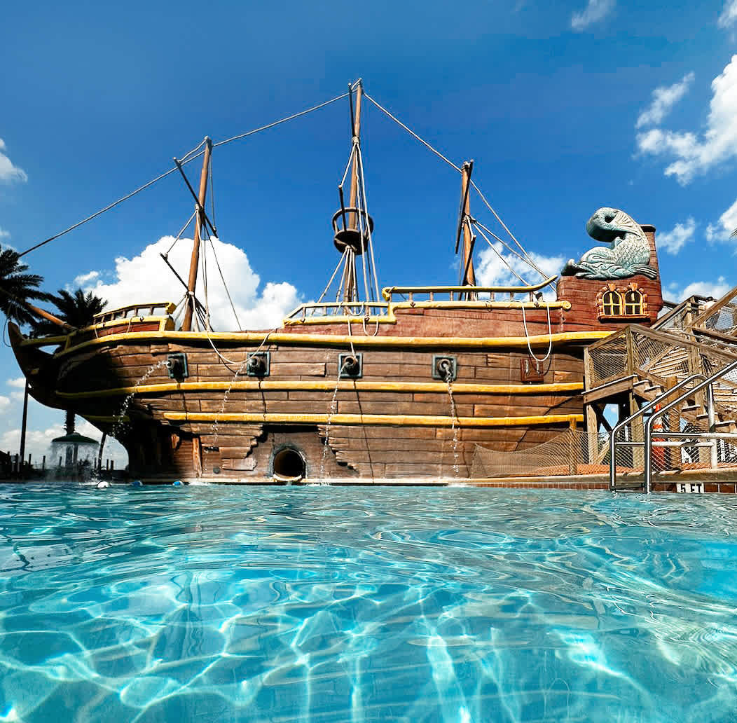 A close-up, low-angle view captures the impressive wooden pirate ship structure within a clear blue swimming pool, showcasing its intricate details and water-splashing features. The bright blue sky with fluffy clouds is visible above.