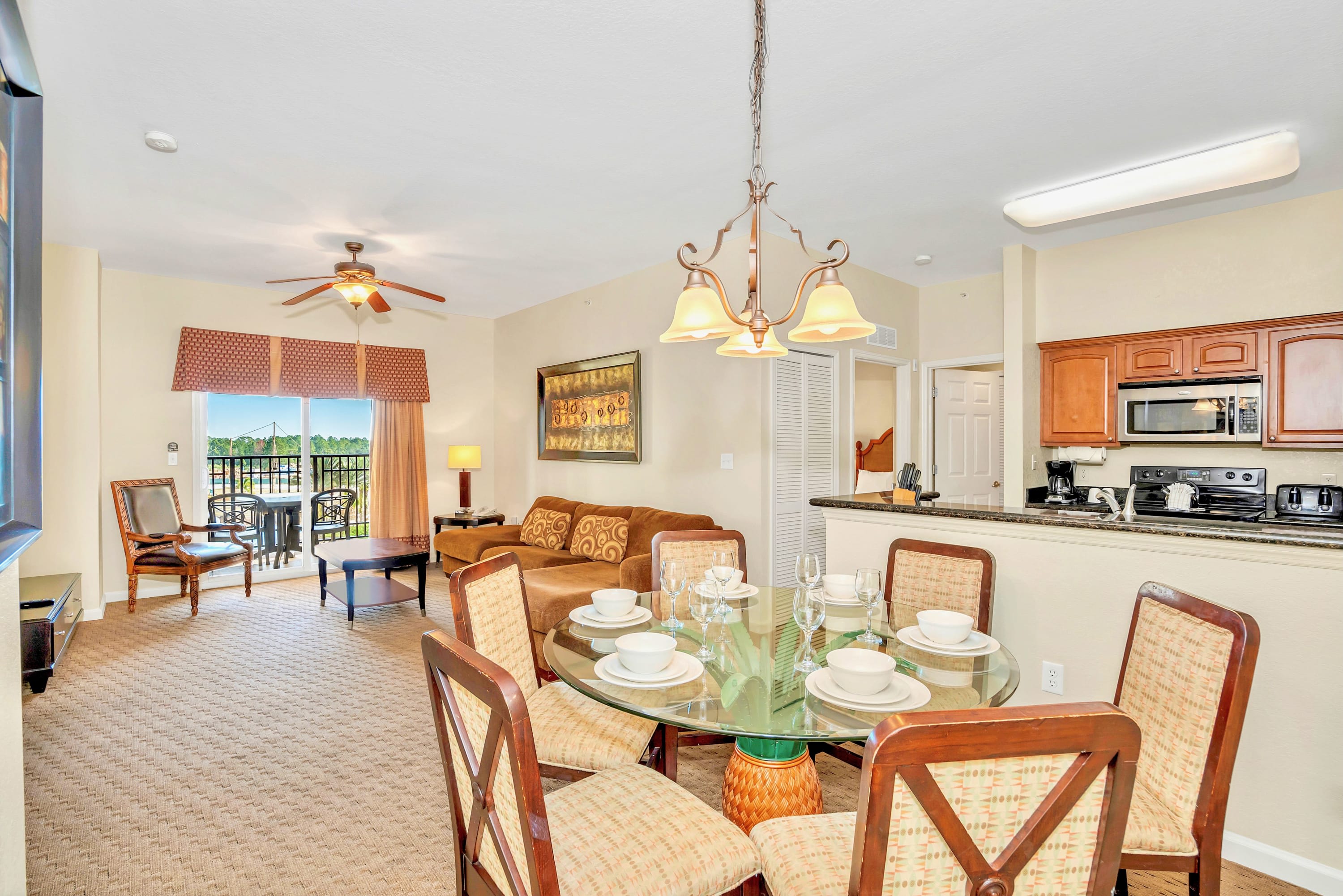 A dining area with a round glass-top table and four chairs with patterned fabric seats is centrally located, with a view of the living room and a compact kitchen. A chandelier hangs above the table.