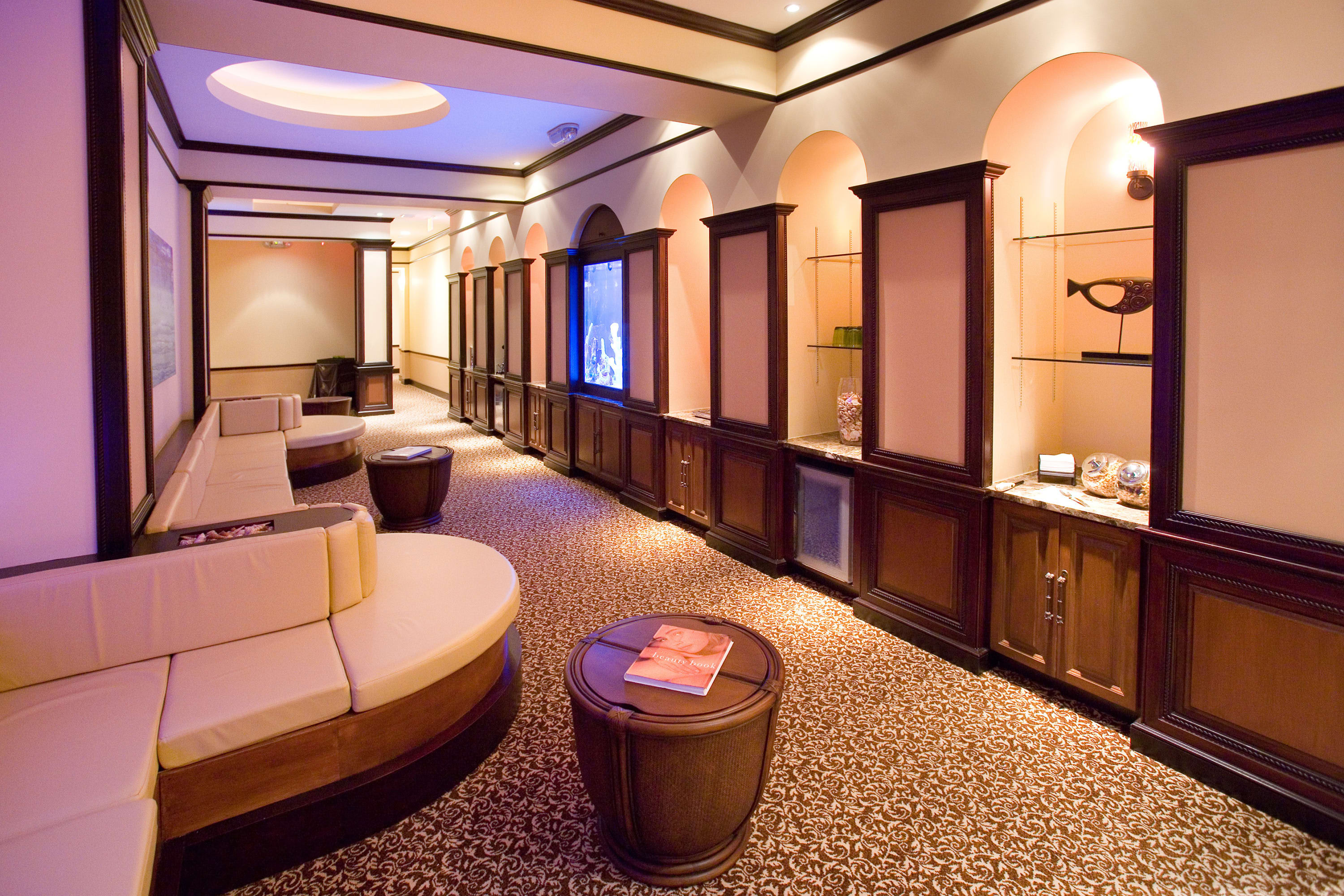 A dimly lit common area features curved beige sofas and dark wood accent tables, set against walls with arched alcoves and built-in shelving. The space is carpeted with a patterned rug.