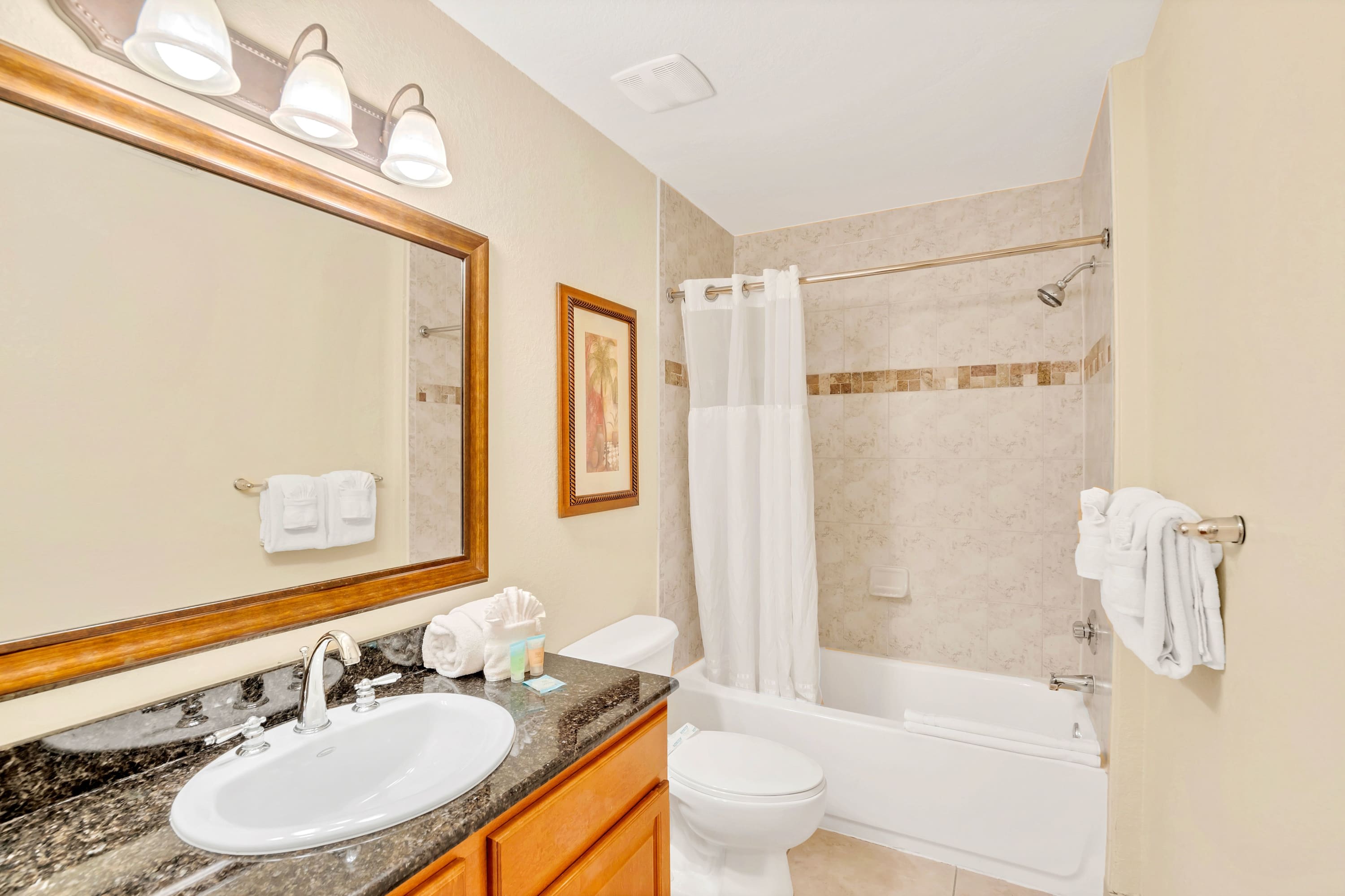 A bright bathroom features a dark granite-look vanity with a white sink and a large wooden-framed mirror. A white shower curtain encloses a tiled tub/shower combo, and a toilet is positioned next to the vanity.